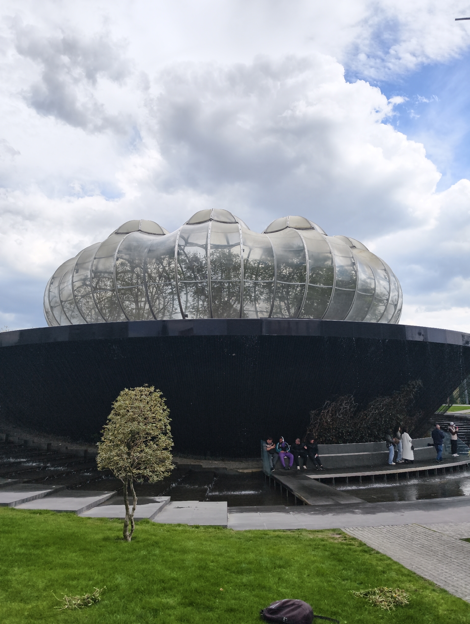 The image shows the "Galitsky Park" in Krasnodar, Russia. A modern architectural structure is prominent, featuring a black, bowl-shaped base with water cascading down the sides, topped by a glass dome with several lobe-like protrusions. Inside the dome, the reflections of trees are visible.
Several people are present: a group is sitting on a bench along the water's edge, while others are standing or walking nearby. There is a small, isolated tree on a grassy patch in front of the structure.
The sky is overcast with numerous puffy white clouds. Paved areas are visible around the fountain and grass. A black backpack is resting on the grass in the foreground.
