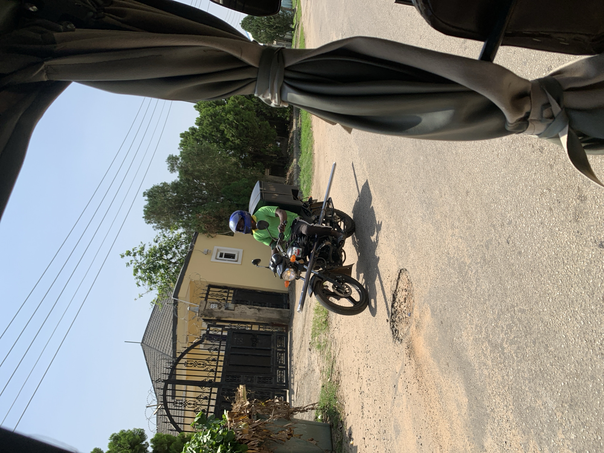The image shows a person riding a motorcycle in what appears to be a residential area. The rider is wearing a green shirt and a blue helmet, and the motorcycle has a black cargo box attached to the back. The setting includes a yellow house with a black metal gate and fence, and several trees. The ground is a paved road with some potholes. Power lines are visible in the sky, suggesting an urban or semi-urban environment. The sunlight indicates it is daytime, likely mid-morning or afternoon, given the clear sky and shadows. The overall scene suggests a person engaged in delivery or transportation. There is no visible text in the image.