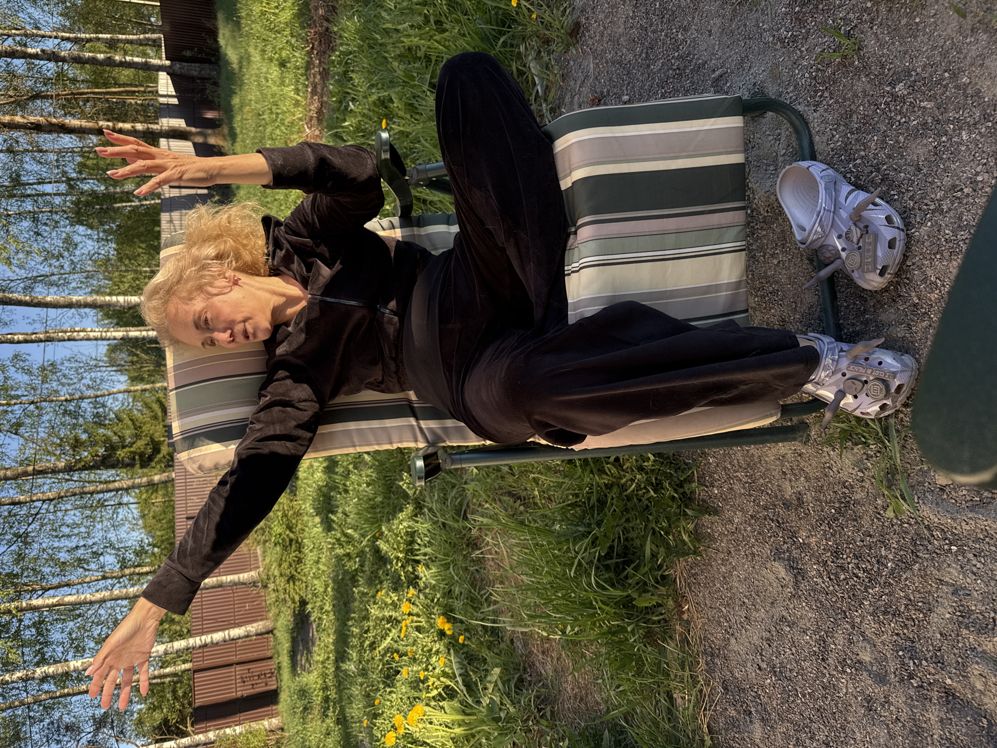 A woman with blonde curly hair, wearing a black velour tracksuit and decorated Crocs shoes, is reclining on a striped lawn chair outdoors. Her arms are extended outward. One of her shoes is next to the chair on a gravelly area. The scene includes a grassy area with dandelions, a wooden fence, and birch trees. The location is outdoors, potentially a yard, near Steklyannyi, Russia.