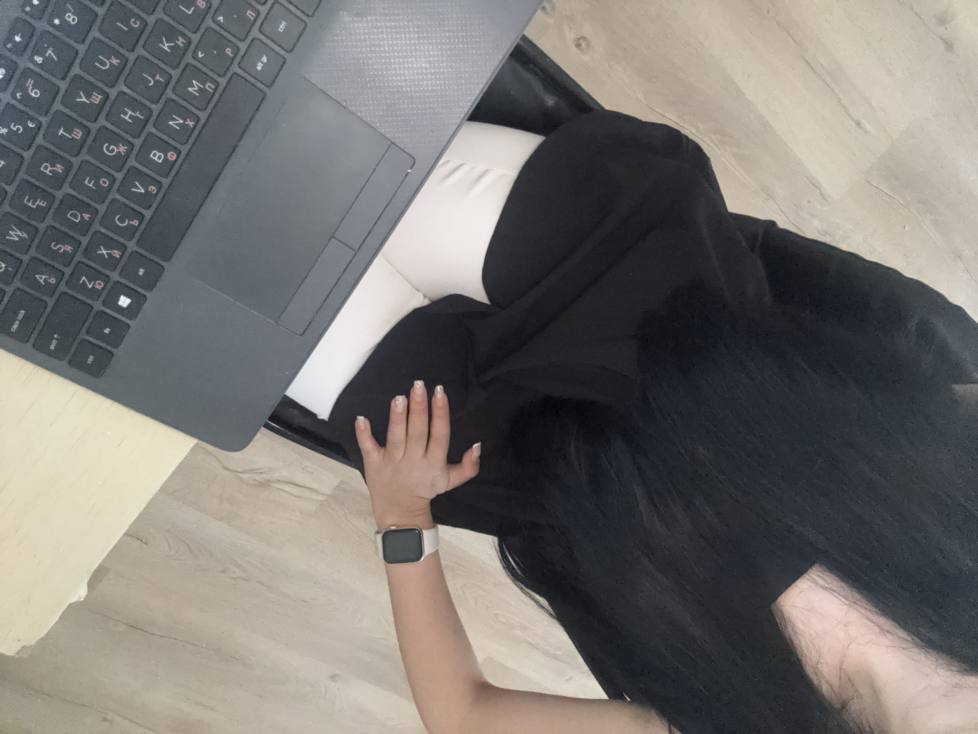 A high-angle shot depicts a person seated at a desk using a laptop. The laptop is open, displaying a keyboard with Cyrillic characters. The person's left arm is visible, adorned with a smartwatch featuring a white band. Their hand rests on a black surface, possibly clothing or a seat. Long, dark hair cascades over the person's shoulder and arm. The person is wearing white pants. The surrounding environment features light-colored wooden flooring and a white desk. The location is unknown, I cannot verify that this image was taken in Plovdiv, Bulgaria.