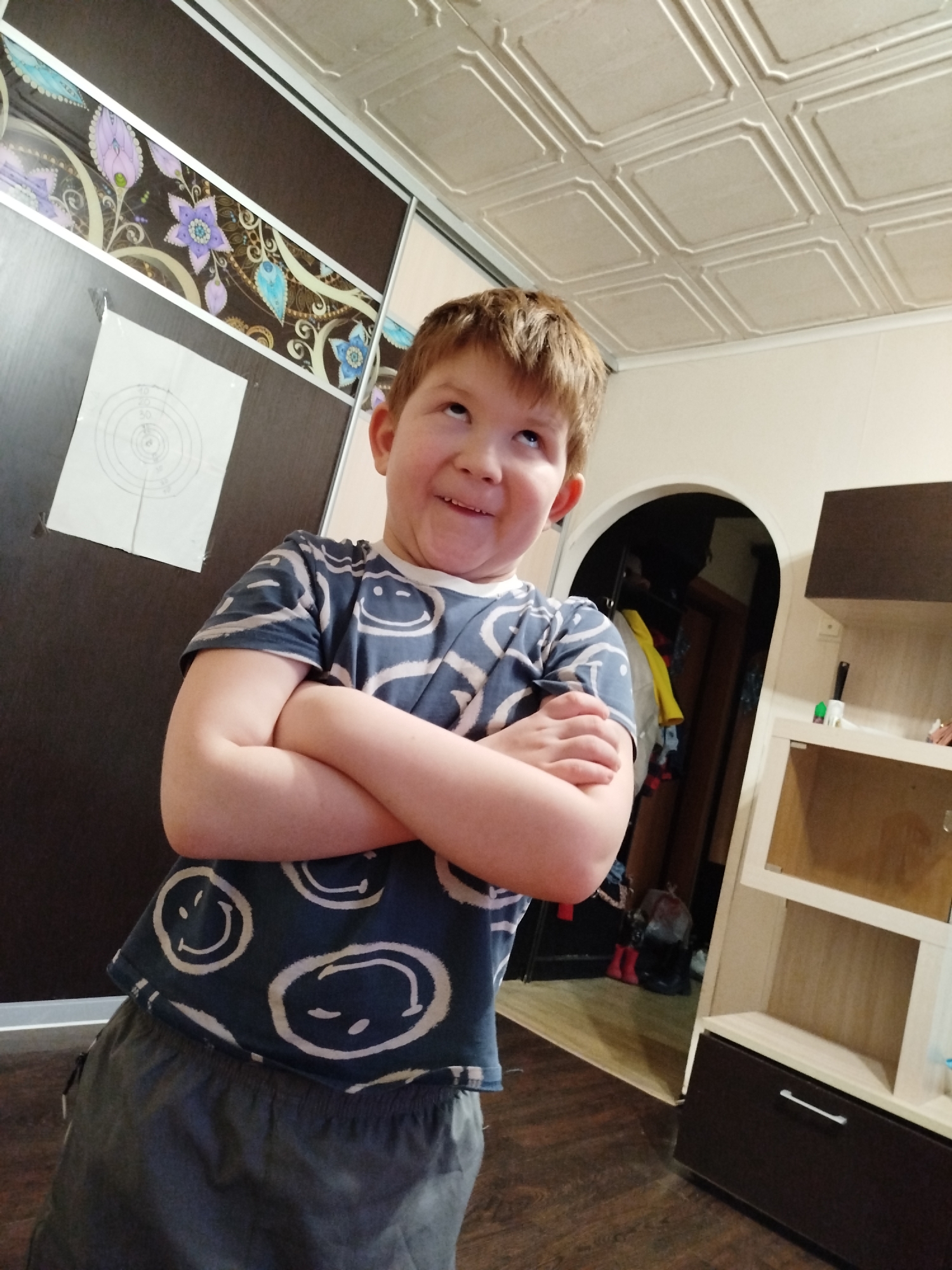 A fair-skinned boy with short brown hair stands indoors, his arms crossed, looking upward with a slight grin. He wears a dark blue t-shirt with white smiley face designs and gray shorts. Behind him to the left is a dark-paneled wardrobe with a floral design at the top and a target pinned to it. To the right is a shelf unit. The background reveals an arched doorway leading to a room with clothes hanging. The ceiling has a patterned, tile-like appearance. The location is in the city of Chaikovsky, Russia. The floor appears to be wooden.