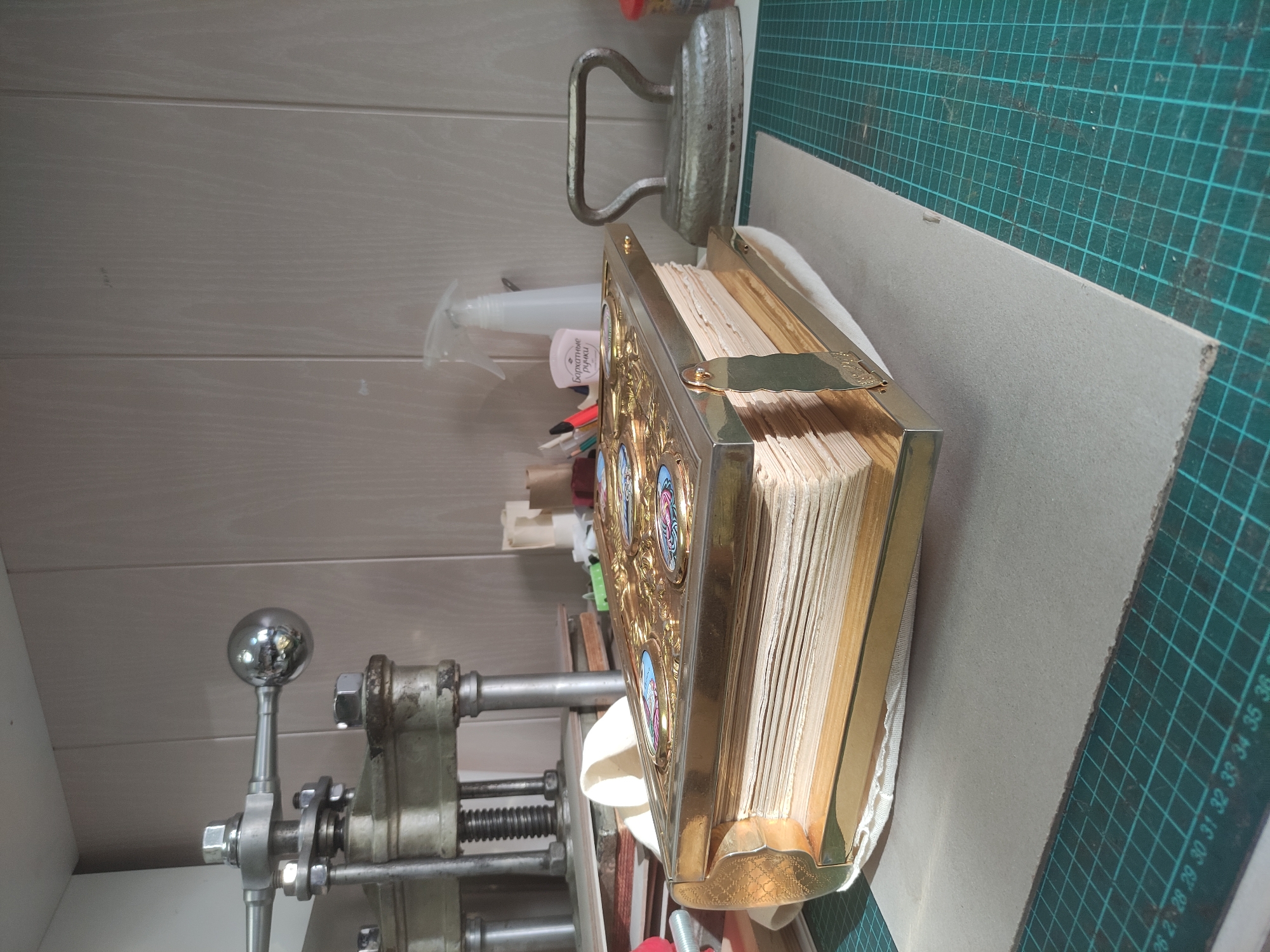 The image depicts a workbench in a bookbinding or restoration workshop located in Moscow, Russia. The central subject is a large, ornately decorated book with a thick, golden-colored metal cover. The cover features intricate embossed designs and multiple oval-shaped inlays displaying colorful iconographic images. The book's pages are thick, aged, and light-colored. It rests on a light grey piece of cardboard, which is padded by white fabric and placed on a green self-healing cutting mat with a white grid and numerical markings.
In the left background, a heavy, silver-colored metal book press is visible, featuring a large spherical handle, threaded rods, nuts, and bolts. It sits on stacked wooden and paper materials.
In the right background, an antique cast-iron flat iron is present. Also on the workbench are a clear plastic spray bottle with a white nozzle and a pink container labeled "Бархатные ручки" (Velvet Hands). Additional workshop items, including pens, pencils, and spools of thread, are visible. The background wall is composed of light-colored, vertically paneled material. No individuals are present in the scene.