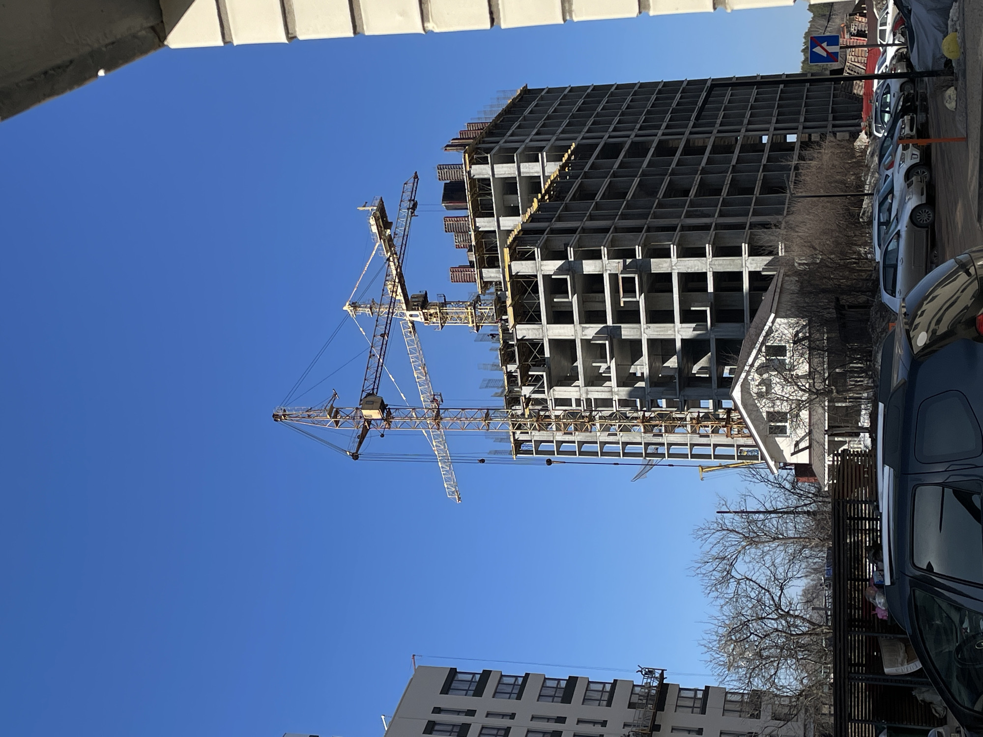 The image shows a construction site in Izhevsk, Russia. A high-rise building is under construction, featuring a concrete framework with multiple floors. Two large yellow construction cranes are positioned to assist with the building process. A completed apartment building is visible in the lower portion of the frame. The sky is clear and blue. Parked cars and bare trees are seen on the right side of the image, indicating a street-level view.