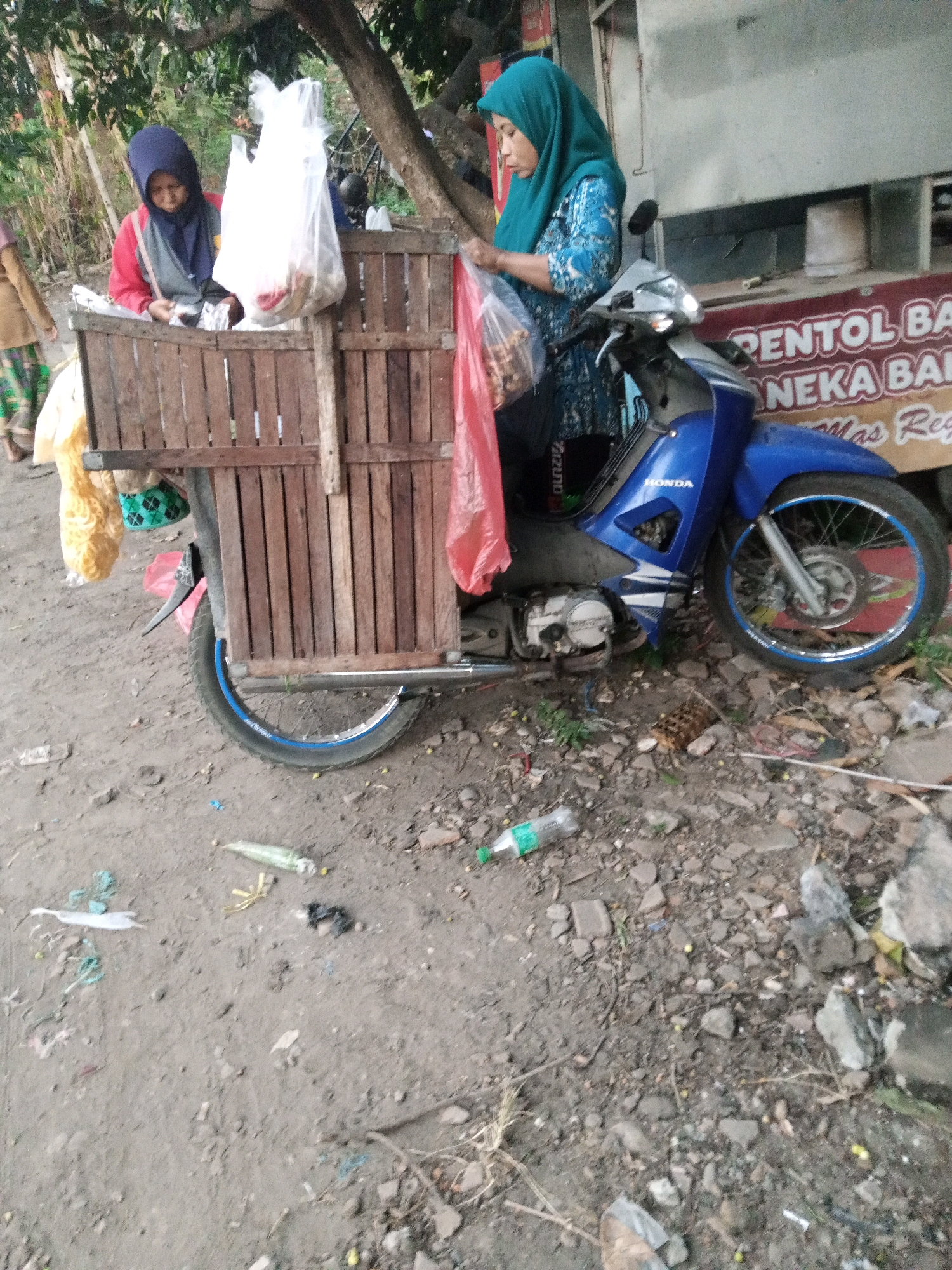An outdoor scene in Sono Ageng, Indonesia, features a blue Honda motorcycle with a wooden cargo box attached to the rear. Two women are near the motorcycle, and a third person is partially visible on the left. The woman on the right, wearing a turquoise hijab and a patterned blue top, is holding a plastic bag of goods. The woman on the left, wearing a dark blue hijab and a red and gray jacket, is looking down at something in the wooden box. The motorcycle is parked on a dirt and gravel surface littered with debris, including plastic bottles and trash. In the background, a building with a sign that reads "PENTOL BA NEKA BA MAS Reg." is visible. Trees and foliage are present in the upper portion of the image.