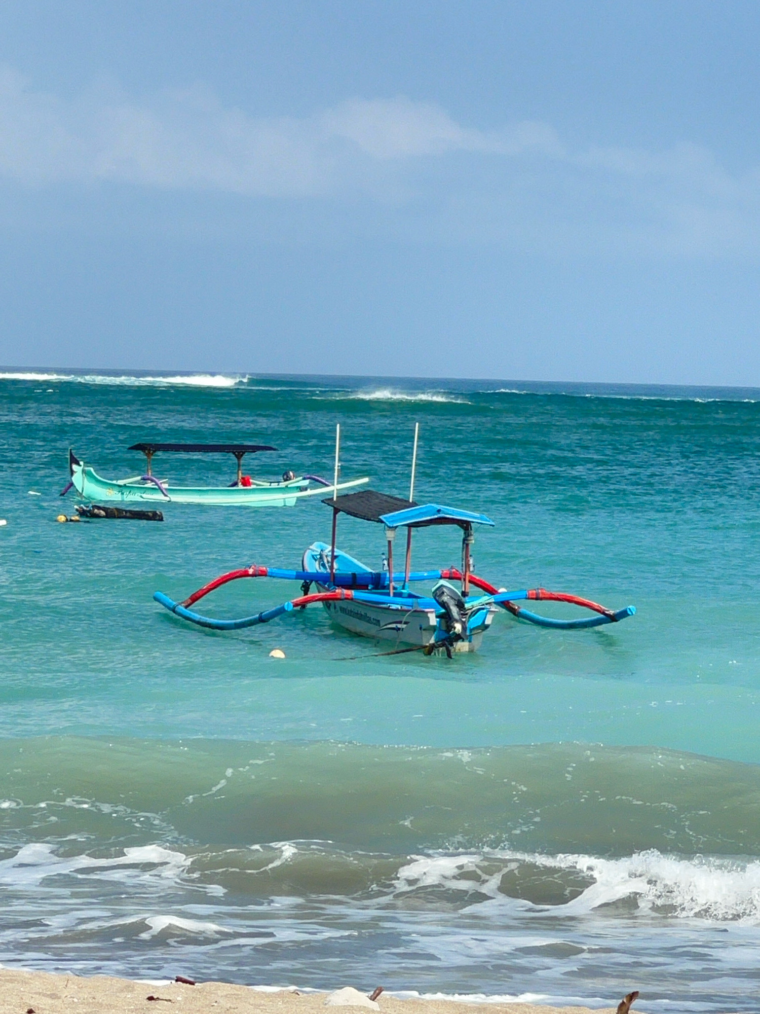 Two Indonesian jukung boats are moored in the turquoise waters off Kuta Beach, Bali, Indonesia. The boat in the foreground is blue and white with a canopy and an outboard motor. It has two distinctive outriggers extending from its sides, painted red and blue. The second jukung, further offshore, is a pale green color with a similar canopy and outriggers. The water is calm, with gentle waves approaching the shore where sand and some driftwood are visible. The sky is clear and blue with a few scattered white clouds. There are no people visible in the image.