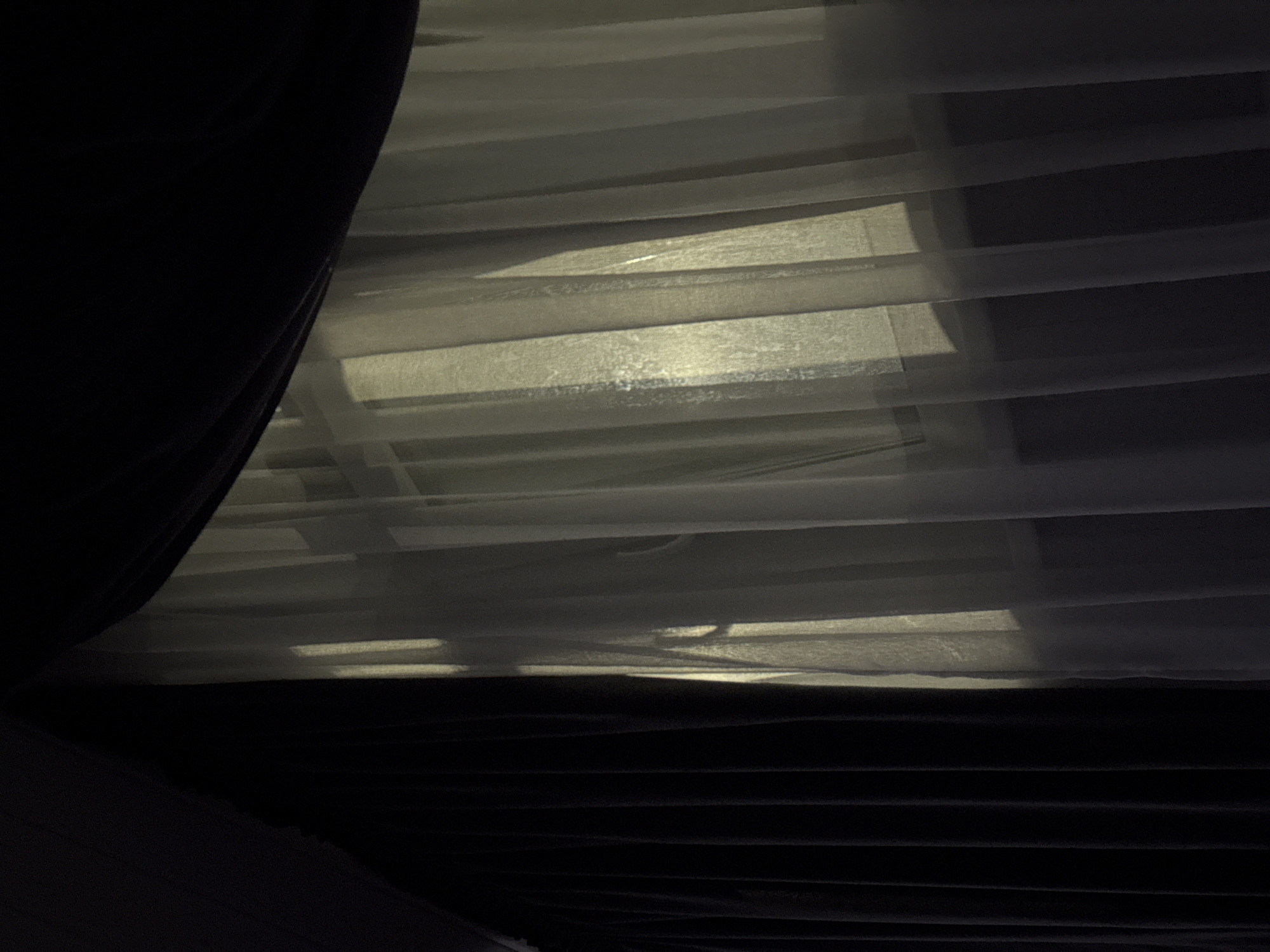 The image shows a close-up view of dark curtains on the left and sheer white curtains on the right, allowing light to filter through from behind. Vertical folds are visible in both sets of curtains. The lighting suggests it is either dawn or dusk, with soft, diffused sunlight casting subtle shadows through the sheer fabric. The environment appears to be indoors, possibly a residential setting, given the presence of curtains. There are no people or overt actions depicted in the scene. The location provided, "Doma 1096 Km, Russia," does not offer any visual cues within the image itself.