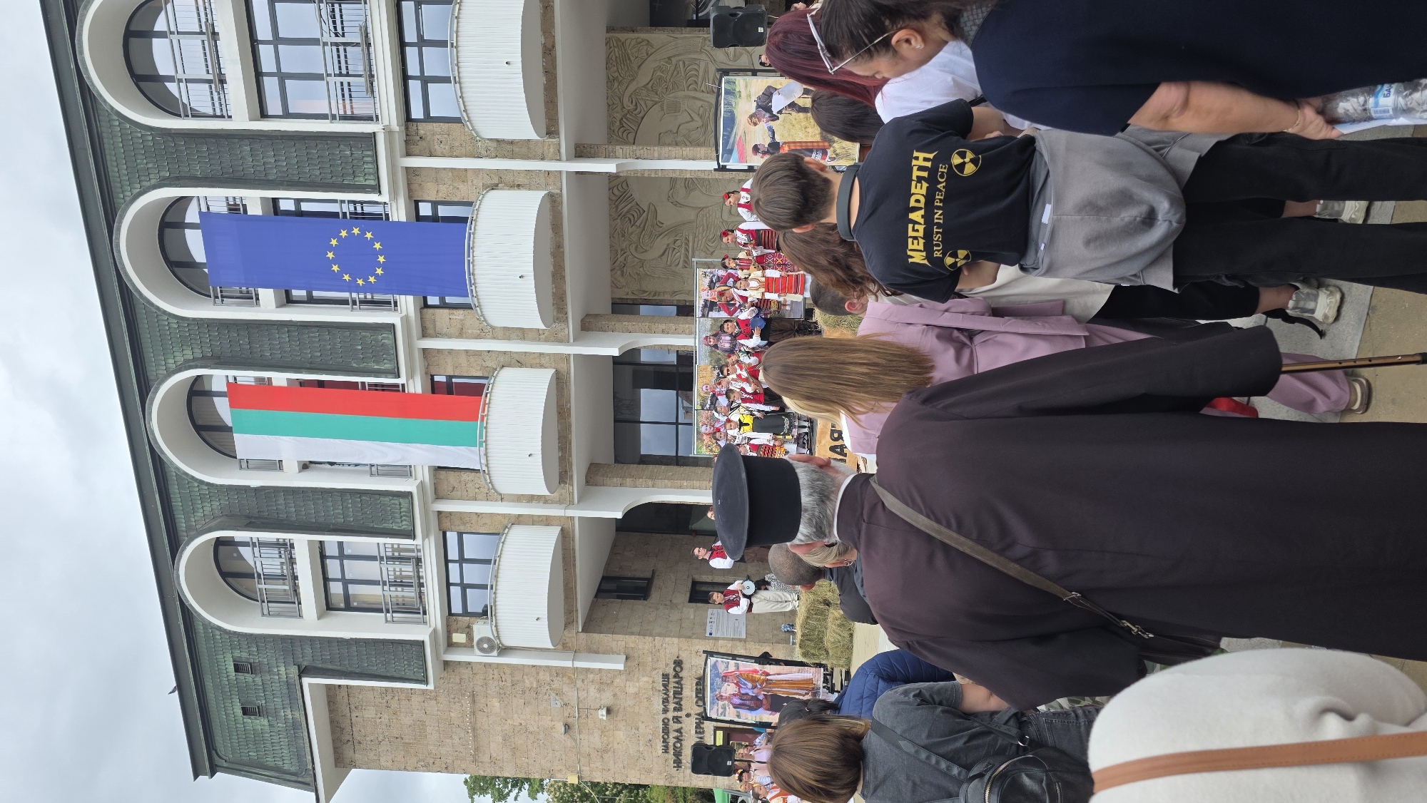 The image shows a crowd of people gathered in front of a building. Two flags, the European Union and Bulgarian flags, hang from the building's balconies. Individuals in traditional folk costumes are visible on a stage. The crowd is composed of people of varying ages, with some looking at the stage. A man in a black shirt with "Megadeth Rust in Peace" printed on it is visible. A man in a black hat is also visible. The building's facade is beige with arched windows and balconies. Text in Bulgarian, "НАРОДНО ЧИТАЛИЩЕ НИКОЛА И. ВАПЦАРОВ-ПЕРНА ОПЕРА," is visible on the building. This indicates the scene is in Blagoevgrad, Bulgaria, specifically at the Nikola Vaptsarov Community Center.
