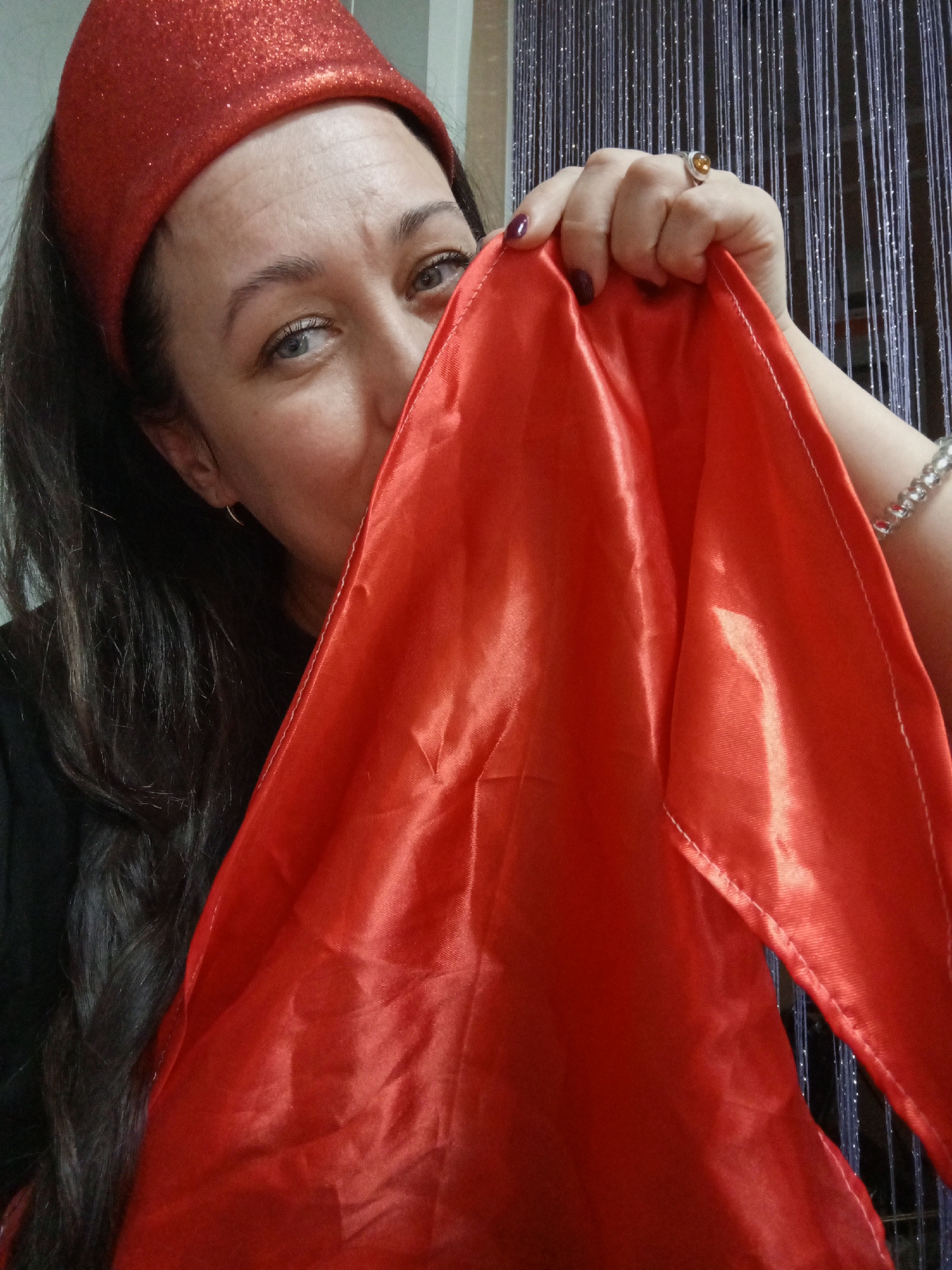 A close-up, indoor portrait features a woman playfully holding a vibrant red, shiny fabric, partially obscuring her face. She looks directly at the camera with a curious expression, her blue-grey eyes visible above the fabric. Her long, dark hair frames her face, and she wears a black top. On her head is a sparkly red headband or hat.
Her left hand, with dark nail polish, holds the red fabric. A silver ring with a prominent yellow or amber stone adorns her ring finger, and a silver beaded bracelet is visible on her wrist. A small, simple hoop earring is seen in her left ear. The background consists of a dark, shimmering curtain or decorative hanging, suggesting an indoor setting in Chaikovsky, Russia. The red fabric appears to be satin or a similar material, with visible white stitching along its edges. The lighting emphasizes the sheen of the fabric and the glitter on her headwear, creating a lighthearted mood. There is no visible text.