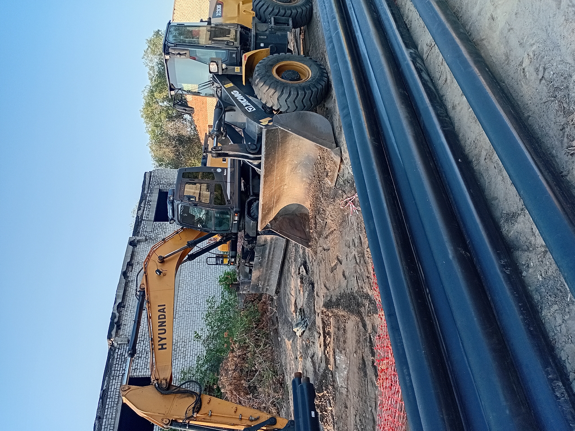 The image depicts a construction site in the city of Semey, Kazakhstan. Two heavy construction vehicles, a yellow Hyundai excavator and a yellow XCMG wheel loader, are parked on a dirt and gravel surface. The excavator's boom is extended, and its bucket is resting on the ground. The wheel loader has its bucket lowered. In the foreground, several large, black pipes are laid out on the ground, alongside a section of orange construction netting. A partially constructed or dilapidated brick building stands in the background, with trees and other buildings visible beyond it. The sky is clear and blue, indicating daytime. The scene suggests ongoing construction or infrastructure work.