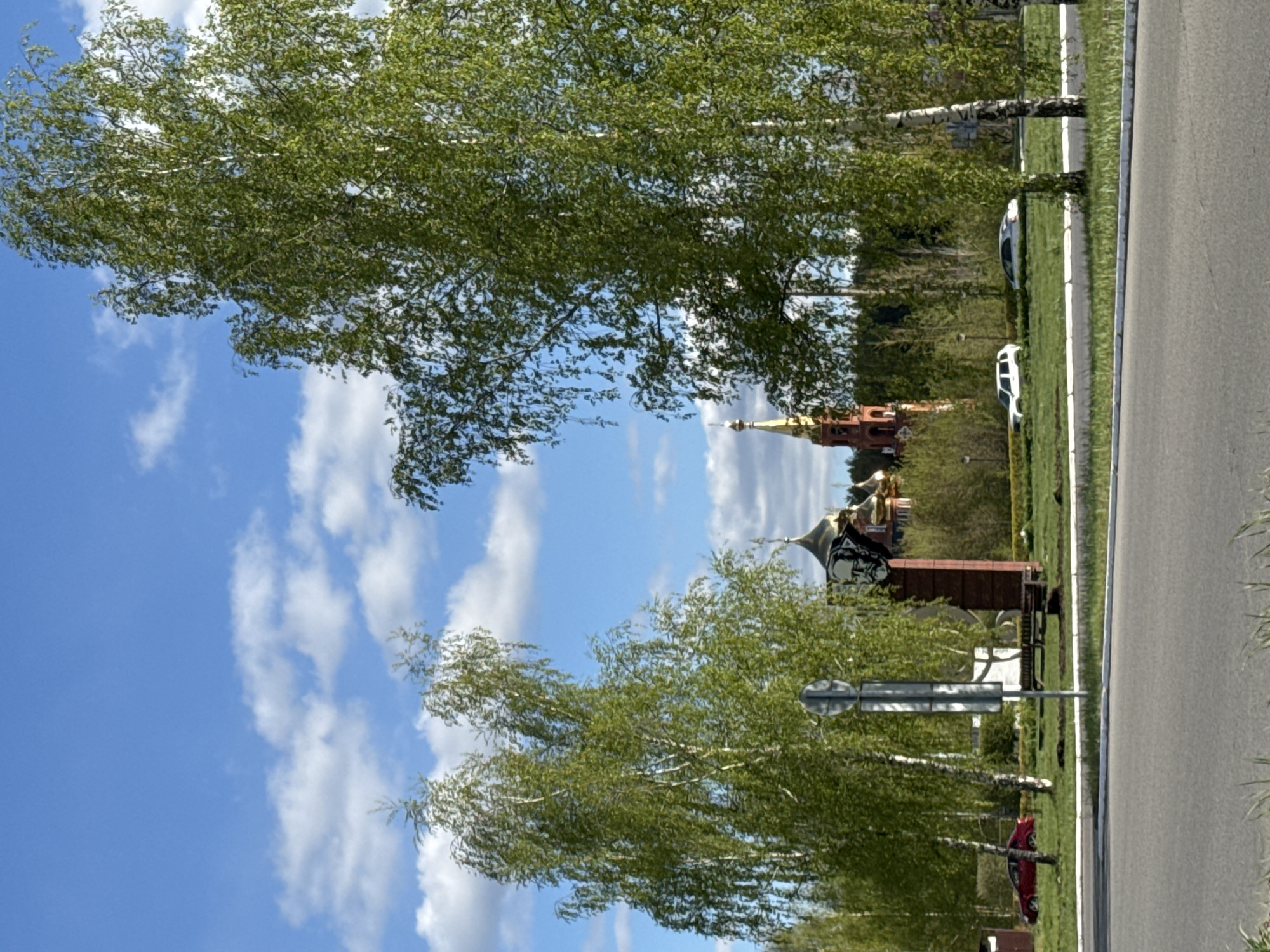 The image depicts a partially obscured view of the Trinity Cathedral in Chaikovsky, Russia. The cathedral's golden domes and red brick structure are visible through the foliage of trees lining a nearby road. A blue sky with scattered white clouds forms the backdrop. A two-lane road runs along the right side of the frame, bordered by a grassy area. Two cars, one white and one silver, are parked on the grass. A red car and road signs are situated closer to the foreground. The focus is on the architectural landmark framed by the natural landscape.