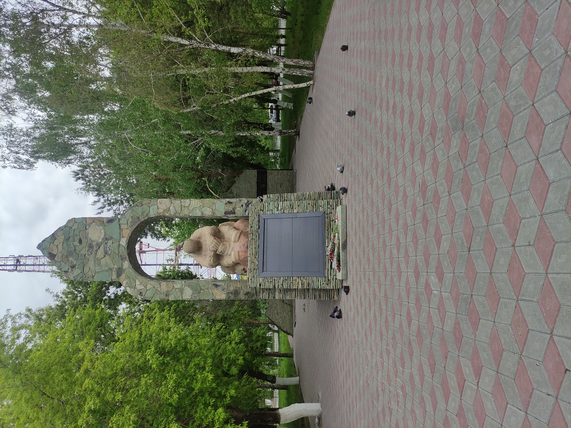The image shows a monument in Pavlodar, Kazakhstan. The monument has a stone arch framing a sculpture of a person in a seated position, with a stone slab adjacent to it. Flowers are arranged around the base of the slab. The area surrounding the monument is paved with patterned tiles, and there are trees and greenery visible in the background. Several pigeons are standing or walking on the paved area. A construction crane is visible through the archway.