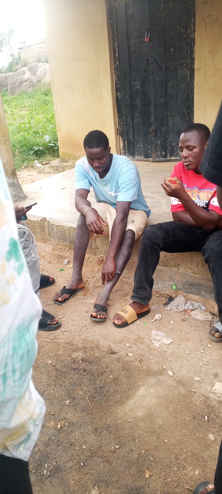Three young men are sitting on a concrete step outside a building with a dark metal door. The man on the left is wearing a light blue t-shirt and light-colored shorts. The man in the middle is wearing a red t-shirt and black pants. He is holding something red in his hands. The man on the right is wearing a black t-shirt and black pants. The ground in front of them is dirt and debris. The building appears to be made of concrete blocks and has a light-colored wall. There is green foliage and rocks in the background.