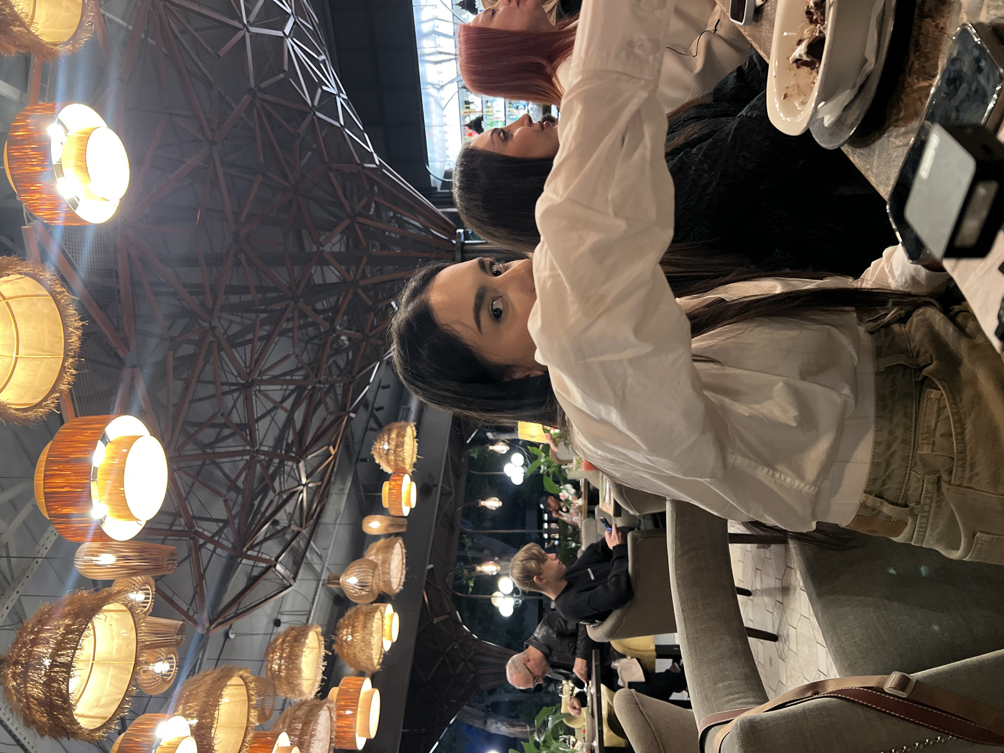 The image captures an indoor scene at a restaurant, likely located in Yerevan, Armenia, given the architectural style and ambience. The setting features a high ceiling with a complex geometric metal framework overlaid with numerous pendant lights that illuminate the space with a warm glow.
In the foreground, there are three women at a table. One with red hair, one with black hair and a third with dark brown hair and fair skin, wearing a white long-sleeve shirt and beige pants, directly faces the camera. Plates, cutlery, and electronic devices are on the table, suggesting a meal or gathering taking place.
In the background, additional patrons are visible seated at tables, indicating a busy restaurant atmosphere. The overall composition conveys a sense of contemporary dining within a stylishly lit interior.