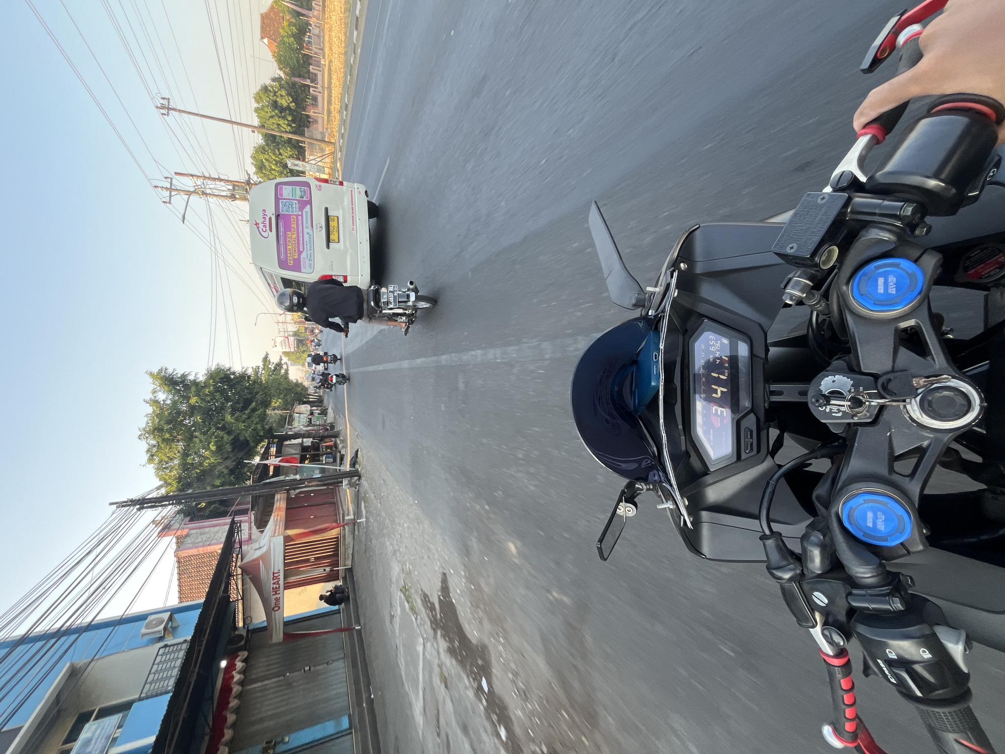 A first-person perspective shot captures a motorcycle rider moving through the streets of Margoyoso, Indonesia, under a clear sky. The road ahead is busy with traffic, including several other motorcycles and a white passenger van labeled "Cahaya" with Indonesian text and advertisements. The motorcycle's dashboard displays "34 km/h" and "6.53". On the left side of the road, a blue building with several closed storefronts and awnings is visible. A person is seated on the curb in front of one of the stores. The right side of the road features a sidewalk, vegetation, and more buildings in the distance. Overhead, a network of power lines crisscrosses the sky.