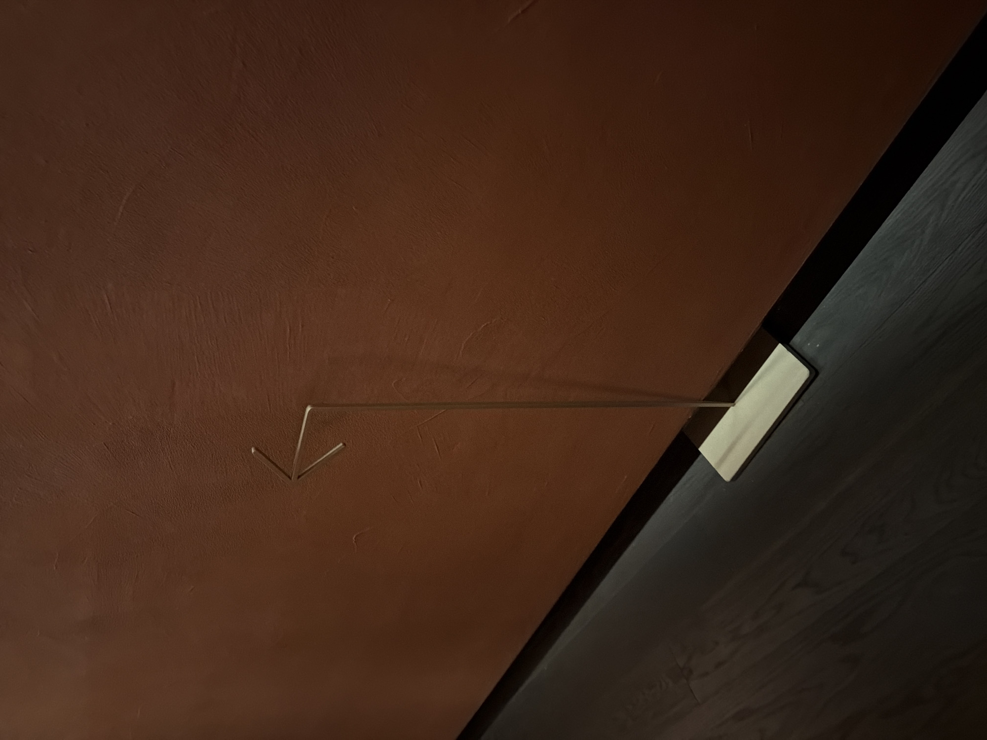 The image depicts a minimalist interior scene with a dark wooden floor and a textured, reddish-brown wall. A thin, metallic arrow-shaped object stands upright on a rectangular wooden base, positioned on the floor and leaning against the wall. The arrow points to the left. The lighting is dim, casting shadows and highlighting the textured wall. There are no people present in the image. The scene is devoid of any identifying markers of a specific location within Seoul, South Korea.