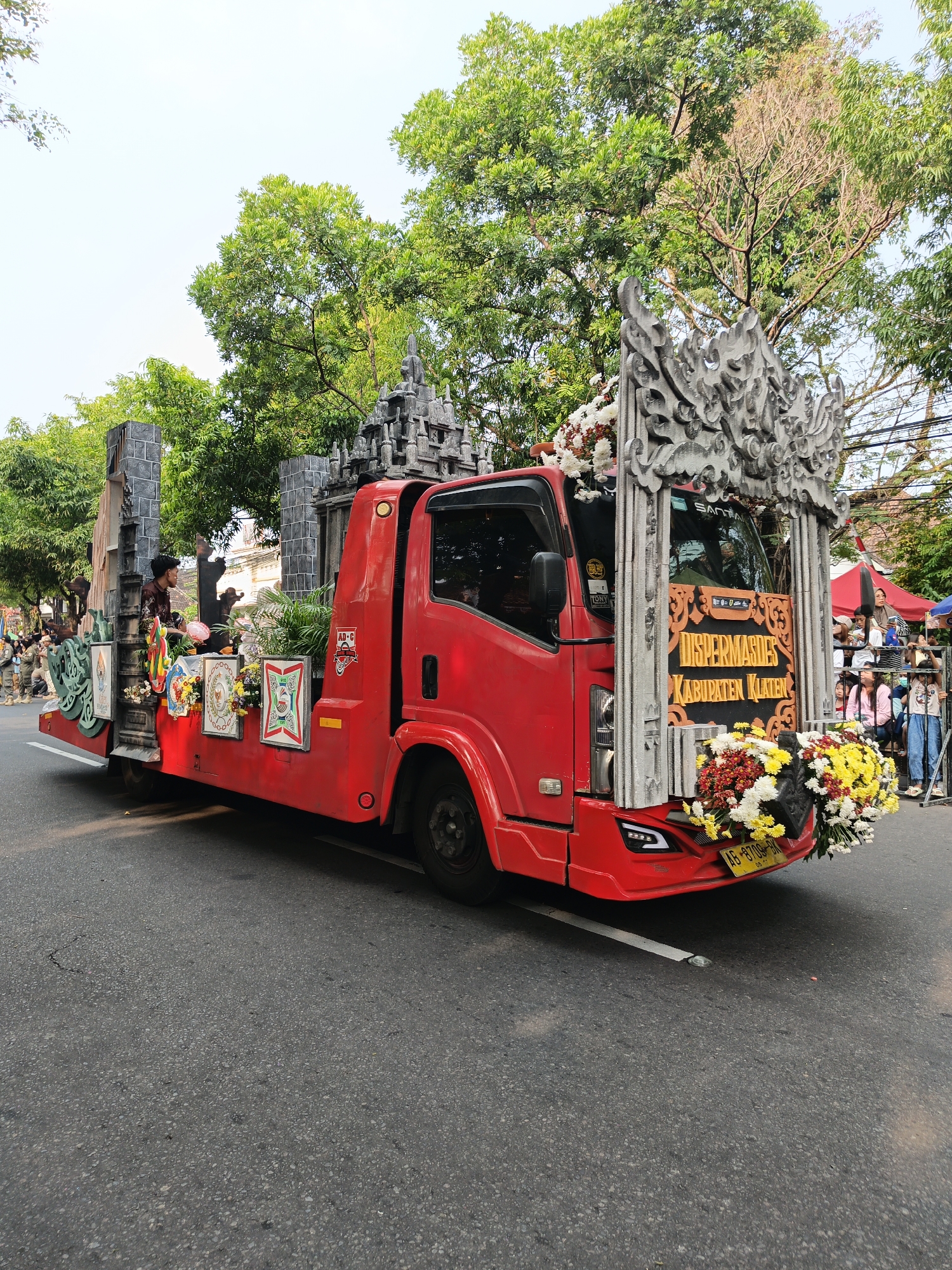 The image captures a bright, sunny day in Gayamprit, Indonesia, with a vibrant red float truck as the central subject. The truck is elaborately decorated with traditional Javanese architectural elements, resembling temple structures, and adorned with abundant floral arrangements in shades of white, yellow, and red. A man in a dark shirt is visible standing on the back of the float, interacting with the decorations. On the side of the float, there are several framed emblems or insignias. The truck is parked on a paved road, with a crowd of onlookers, including children and adults, gathered on the right side of the frame, many of them appearing to be watching the event. Lush green trees with dense foliage line the background, indicating a well-treed urban or suburban environment. The sky is clear and bright. The license plate on the truck reads "AG 8709 BN". The signage on the front of the truck indicates "DISPERMASDES KABUPATEN KLATEN".