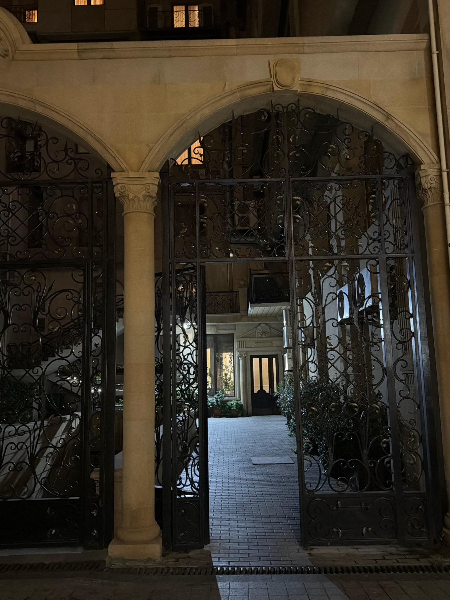 The image shows a view through an arched entryway with ornate black metal gates at night in Baku, Azerbaijan. The entryway leads to a courtyard paved with brick. To the left, through the gate, a staircase is visible, and to the right, dense green bushes are present. In the background, a building with illuminated windows is visible. The architecture suggests an older, possibly residential or historical area. The lighting is dim, characteristic of nighttime, with artificial lights illuminating the courtyard and building windows.