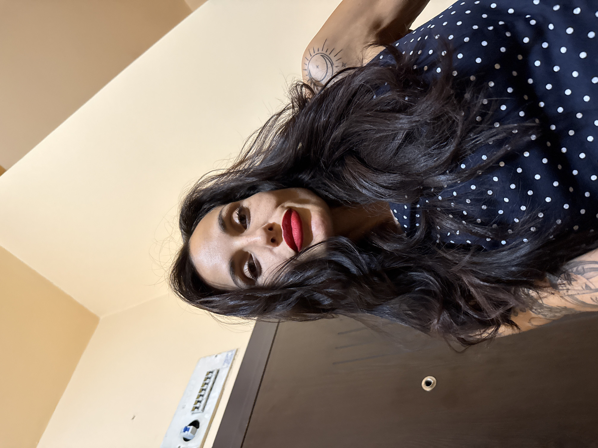 A close-up, low-angle portrait captures a woman with long, dark, wavy hair cascading around her face. She has fair skin, prominent red lipstick, and dark eye makeup, looking directly at the camera with a neutral and intense gaze. She is wearing a dark blue garment patterned with white polka dots. Distinct tattoos are visible on her arms: a sun, moon, and stars motif on her right upper arm, and a partially obscured, larger tattoo on her left arm.
The background consists of light beige-colored walls, indicative of an indoor setting. To the lower left of the frame, a white wall-mounted electrical panel or intercom system with multiple small switches or buttons is visible. Below the woman, a dark brown wooden door or piece of furniture features a small, round metal detail, possibly a peephole or lock. The overall perspective is an upward-looking shot, creating a dynamic and unconventional angle. The scene is brightly lit, likely by indoor artificial light. The image provides no discernible text. This portrait was likely taken in Pleven, Bulgaria.