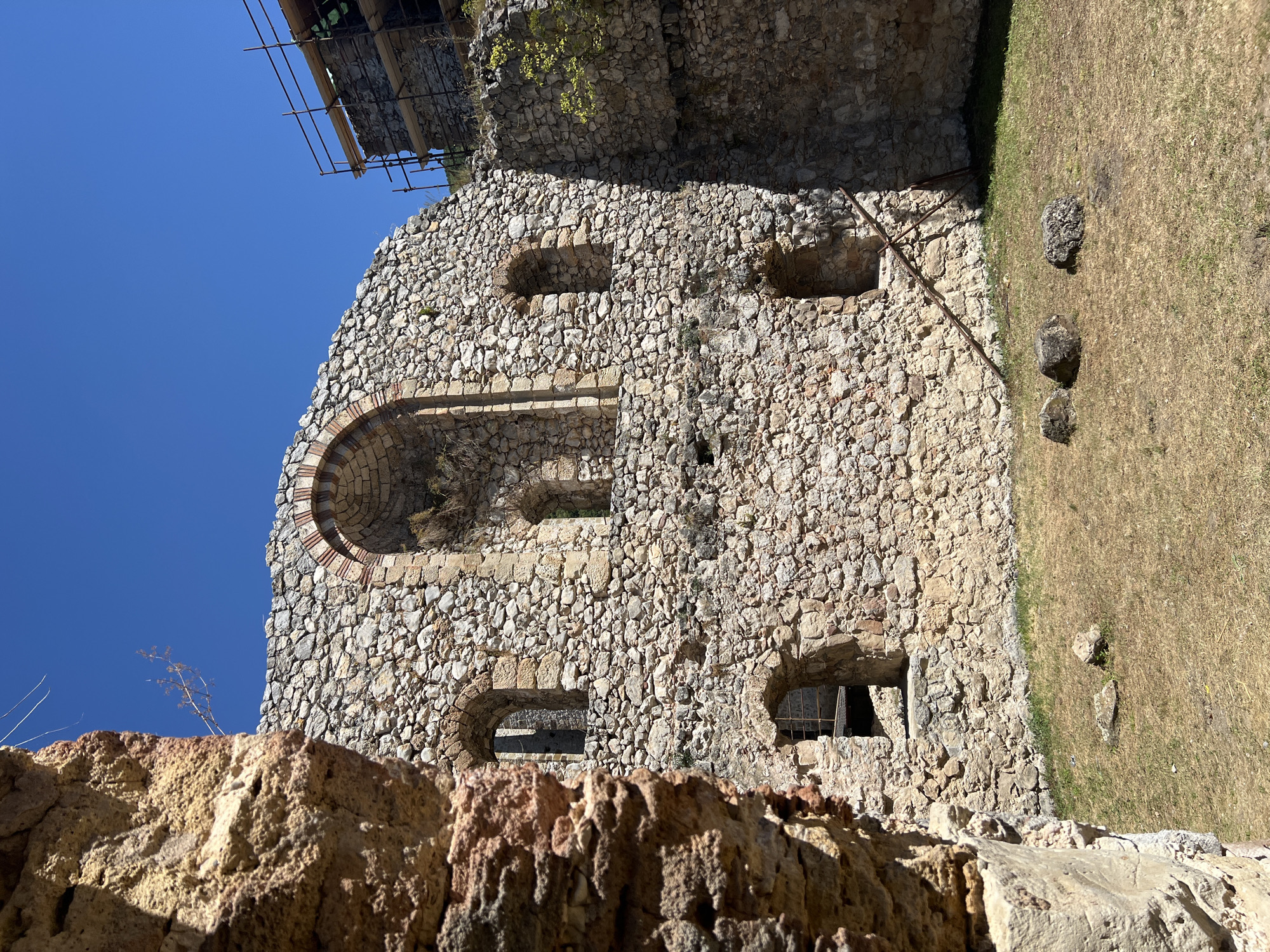 The image depicts the exterior of a stone ruin, likely a historical fortress or castle, under a clear blue sky. The foreground features a weathered, rough-textured stone wall on the left, partially obscuring the view of the main structure. The central part of the image showcases a large, roughly constructed stone wall with multiple arched windows and openings. Some of these openings reveal darkness within, while others offer glimpses of the sky or more stone. A prominent arched opening with reddish-brown bricks is visible near the top center.
The ground in front of the ruin is covered with dry, yellowish grass and a few scattered rocks. To the far right, another section of stone wall is visible, and scaffolding is erected against it, suggesting ongoing renovation or preservation work. A thin, dry branch protrudes from the top of the main stone wall, reaching towards the sky. The overall impression is one of ancient architecture in a state of disrepair, with signs of human intervention for preservation.
This ruin is located in the city of Despotovac Municipality, Serbia, specifically at the Manasija Monastery complex, which is known for its well-preserved medieval fortress.