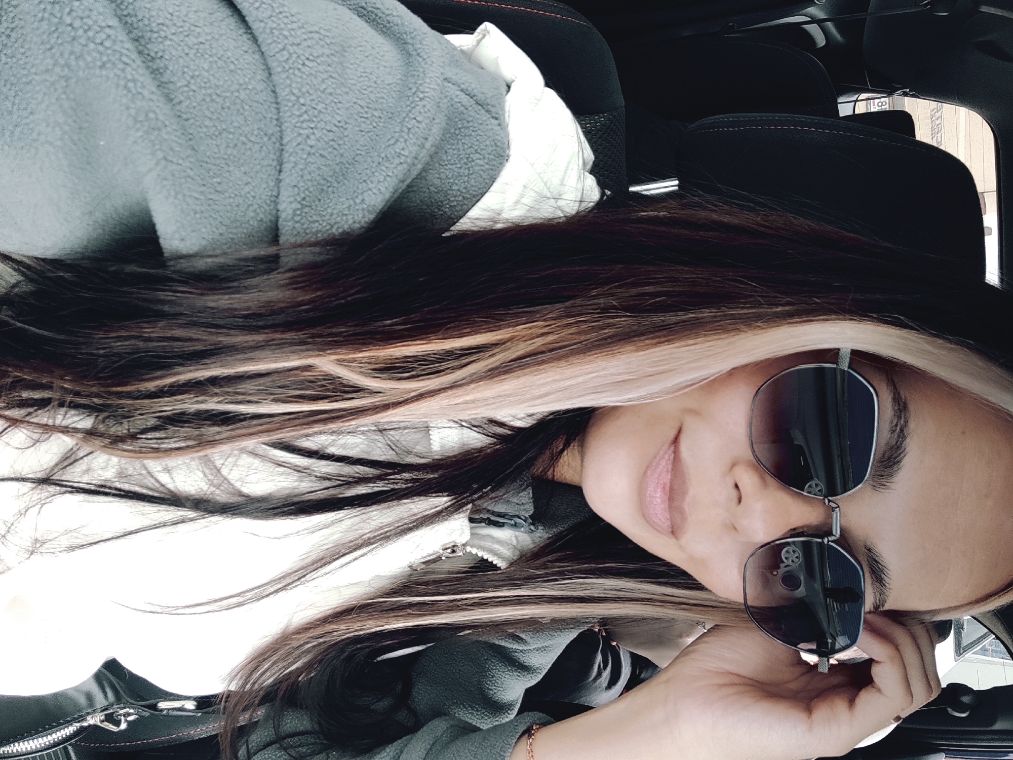 A selfie of a woman in a car. She wears sunglasses and a zippered jacket, with her hair in a dark-to-light ombre style. The car's interior is dark and appears to have leather seats with red stitching. Outside the car window, a building with a partially visible sign reading "8" can be seen. Based on this sign, the location is likely Chaikovsky, Russia, where a business called "8th Floor" exists.