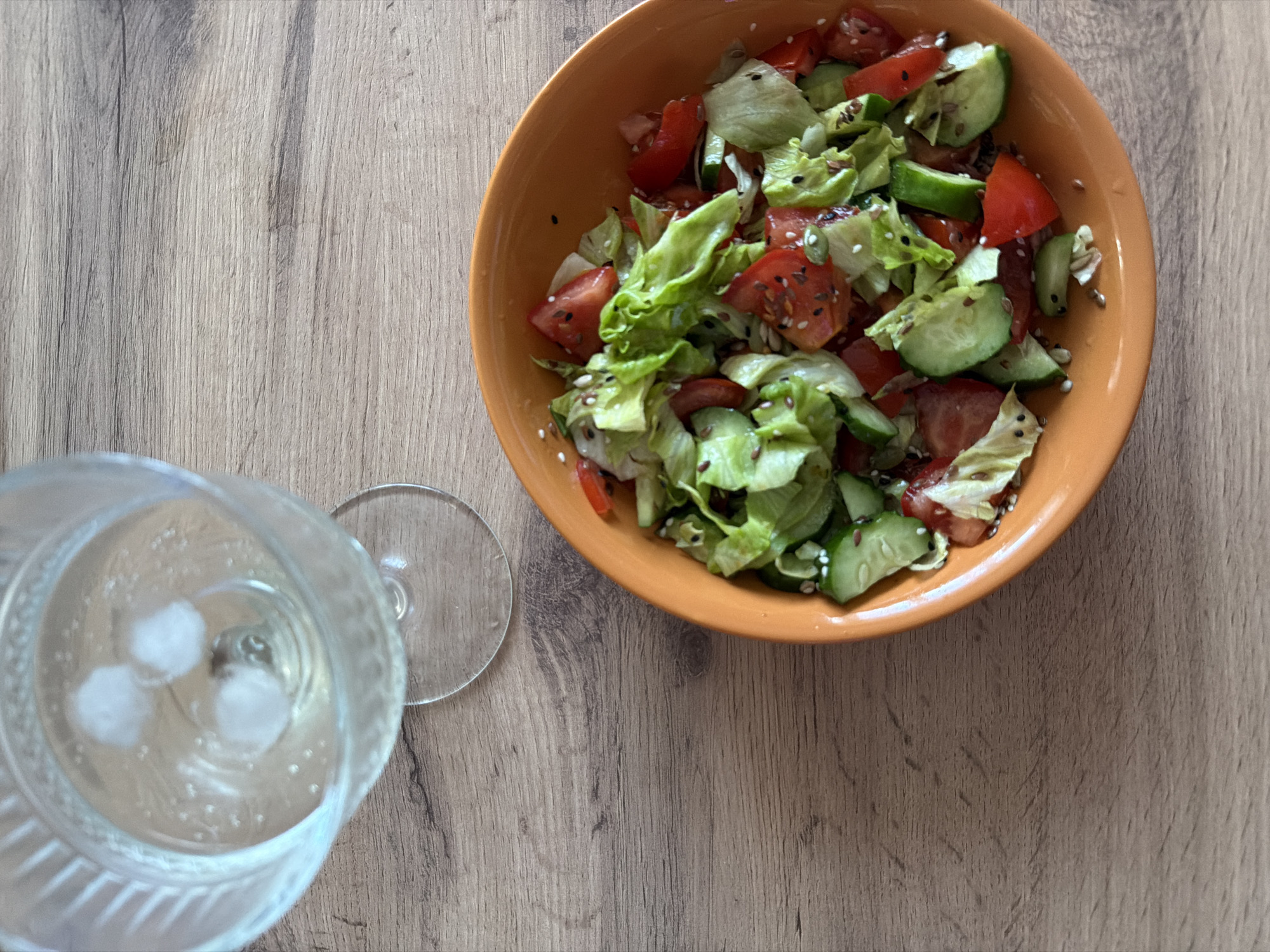 An overhead shot presents a simple meal setting on a light-colored wooden table. A round, orange-terracotta bowl filled with a fresh salad occupies the upper right portion of the frame. The salad consists primarily of vibrant green lettuce, diced red tomatoes, and sliced green cucumbers. It is sprinkled with various seeds, including white sesame seeds, golden flax seeds, and some smaller dark seeds, adding texture and visual interest.
To the left of the bowl, occupying the lower-left corner, is a clear glass, possibly a goblet or a sturdy stemmed glass, holding a transparent liquid with three visible ice cubes. The glass appears to have a textured or ribbed pattern on its lower half. The stem and base of the glass are partially visible, indicating its type. The liquid itself seems to be either water or a clear beverage, with a few small bubbles suggesting it might be carbonated.
The wooden table surface features a natural grain pattern with varying shades of light brown and gray, suggesting a plank or laminate design. There are no people visible in the image, and no discernible activity beyond the static arrangement of the meal. The lighting is soft and even, typical of an indoor setting, but specific time of day or external weather conditions cannot be determined. There is no visible text. The scene conveys a calm, everyday moment of refreshment or a light meal. Although the location context is Chaikovsky, Russia, no specific visual cues within the image directly confirm this.