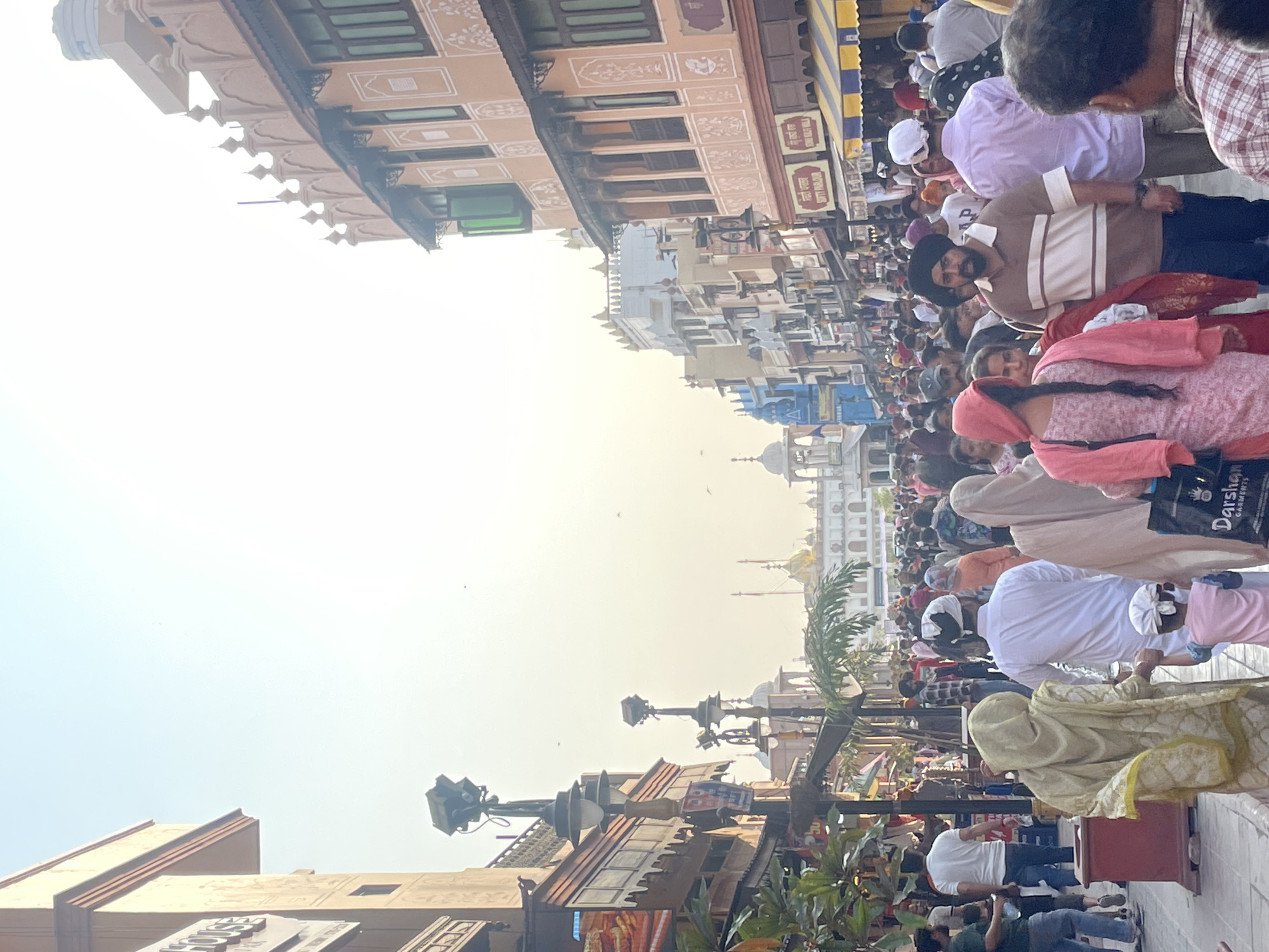 This photograph captures a bustling street scene in Amritsar, India, likely near the Golden Temple. The image is dominated by a dense crowd of people walking along a paved pathway lined with ornate buildings. The architecture is a blend of traditional Indian styles, featuring intricate carvings, arched windows, and domed structures visible in the background. The buildings are painted in warm, earthy tones, with some adorned with decorative patterns. Several lampposts with multiple lights are strategically placed along the street, suggesting it's an area frequented by both locals and tourists. The sky is bright and hazy, indicating a sunny day. The people in the crowd are diverse in their attire, with many men wearing turbans and women in colorful traditional Indian clothing like salwar kameez and saris. Some individuals are carrying shopping bags. The atmosphere appears lively and crowded, typical of a popular pilgrimage or tourist destination.