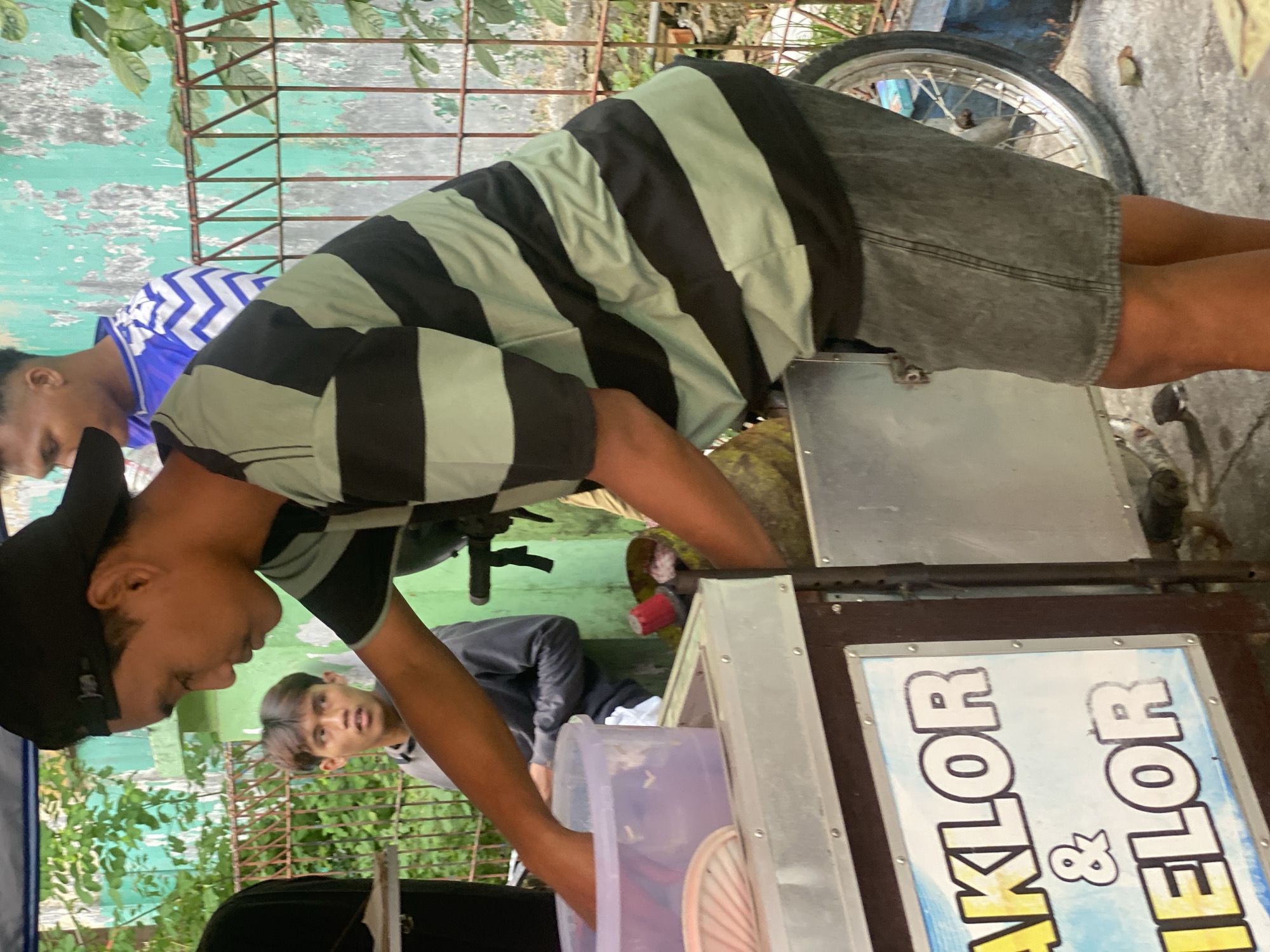 In Banjarbaru, Indonesia, a young man wearing a black baseball cap and a black and light green striped t-shirt is standing next to a food cart. He is reaching into a transparent plastic container on the cart. Behind him, another young man wearing a blue and white chevron patterned shirt is visible. The food cart has a sign that reads "AKLOR & ELOR" in large, bold letters. The cart is positioned in front of a teal-colored, textured wall and a rusty metal fence. Green foliage is present in the background. A bicycle wheel is attached to the side of the food cart. The ground appears to be paved.
