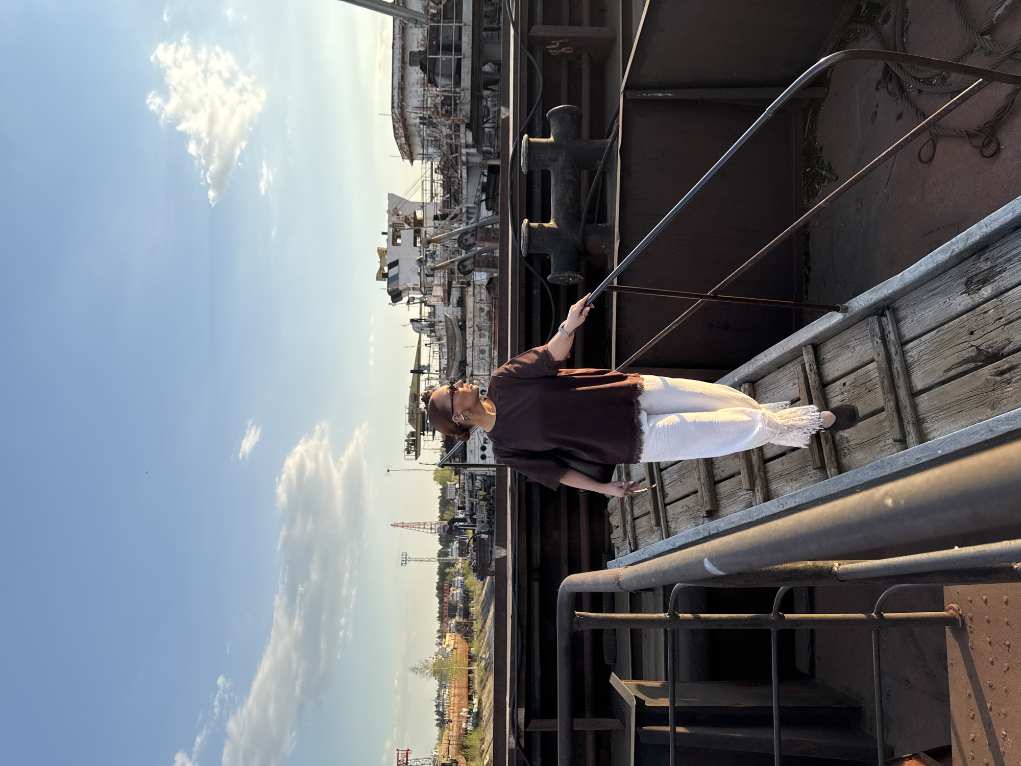 The image depicts a woman standing on a section of a ship or industrial structure, with the cityscape of Chaikovsky, Russia in the background. The woman is wearing a brown top and white pants. She is holding onto a metal bar. The scene is taken from a high angle. The background includes industrial structures, buildings, and a blue sky with white clouds. The lighting indicates it is daytime.