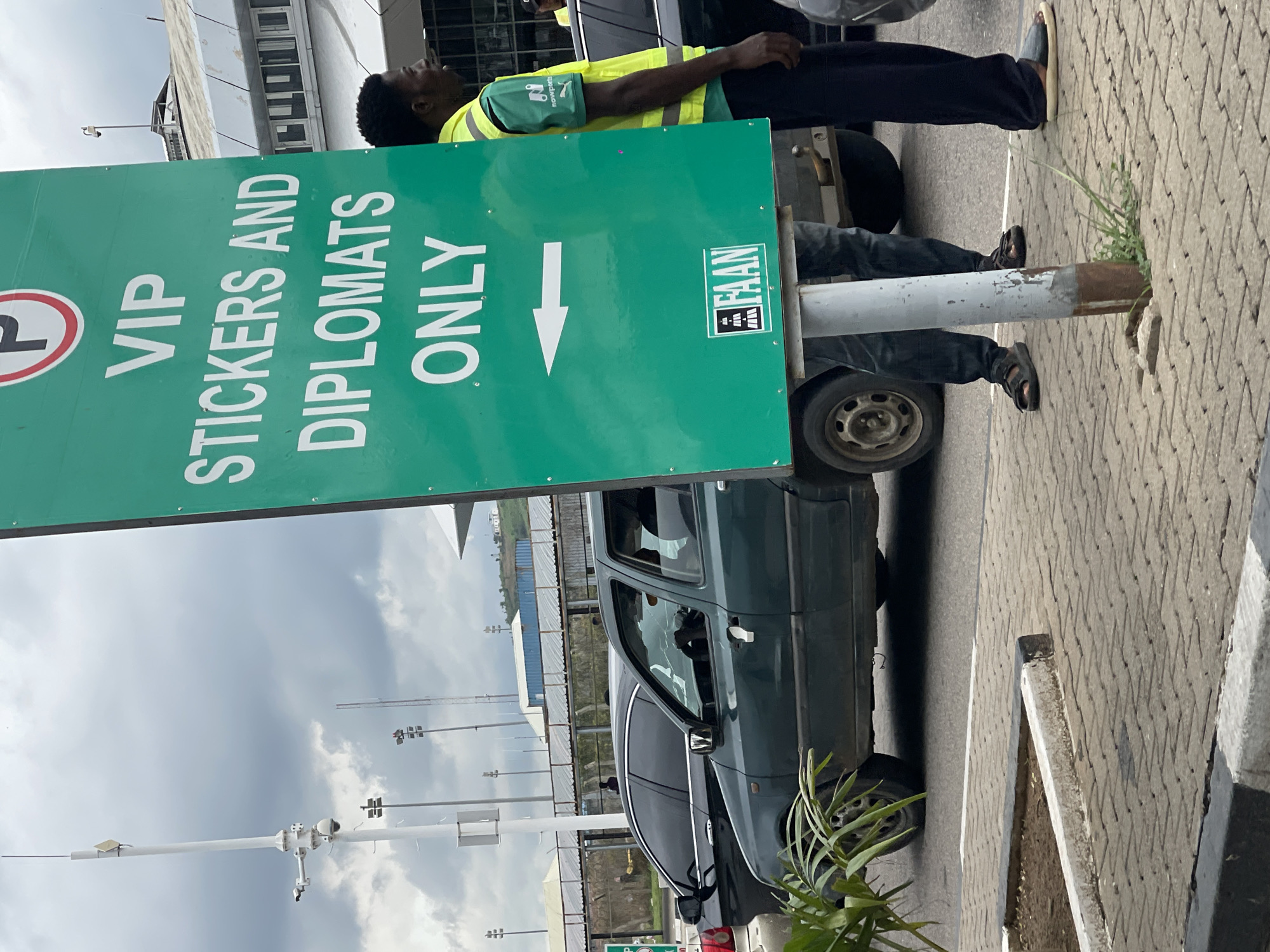The image shows a green sign with white text that reads "VIP STICKERS AND DIPLOMATS ONLY". A white arrow points to the left, indicating a direction. Below the arrow, there is a logo with the letters "FAAN". In the foreground, a man wearing a yellow vest over a green shirt and dark pants is standing next to the sign. Behind him, two cars are visible: a dark-colored sedan and a dark green hatchback. In the background, there are buildings, streetlights, and a cloudy sky. The scene appears to be an airport or a government facility, possibly at Gwaska, Nigeria, given the "FAAN" logo, which stands for Federal Airports Authority of Nigeria. The overall impression is one of an organized entrance or restricted access area.