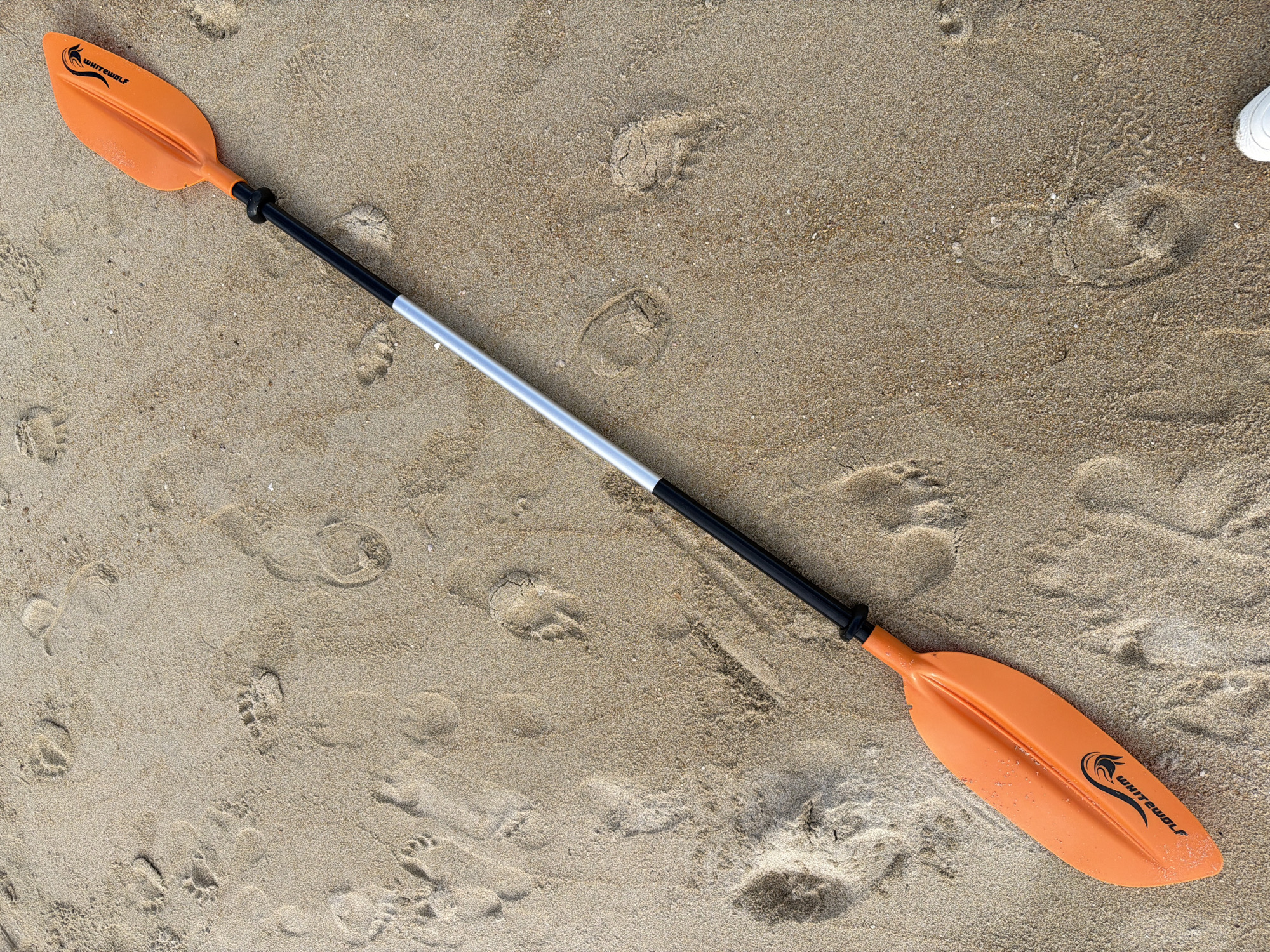 A kayak paddle with two orange blades and a silver and black shaft lies diagonally on sandy ground. The sand is imprinted with various footprints, some appearing to be from human feet and others smaller and possibly animal tracks. The paddle is positioned from the lower left to the upper right of the frame. The background is entirely composed of the sand with footprints.