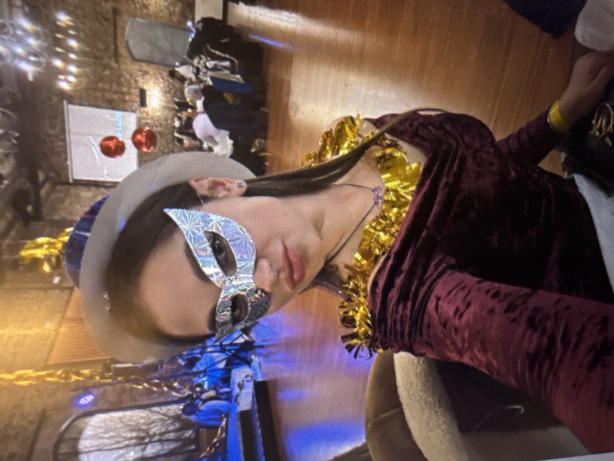 The image shows a medium shot of a young woman wearing a silver masquerade mask, a burgundy velvet dress, and a gold fringe accessory around her neck. She is also wearing a hat with blue and white accents. The background appears to be a dimly lit event space with several tables and chairs set up for a gathering. Red balloons and a projection screen are visible in the background. The lighting is a mix of warm and cool tones, with some areas appearing to be lit by chandeliers or string lights. The floor is a polished wooden surface. It is impossible to determine the location within the city of Murcia, Spain, based on the provided image.
