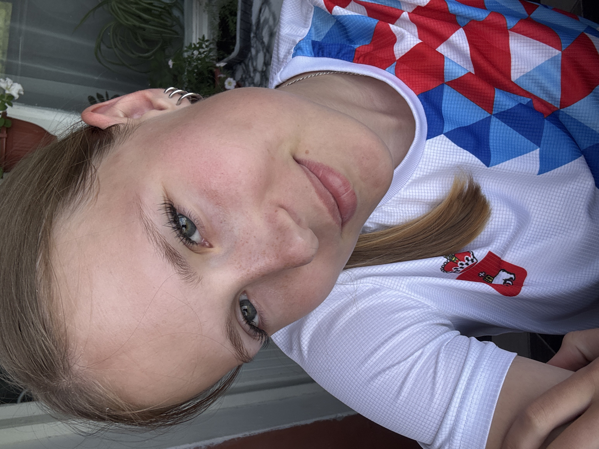 This is a vertical, close-up, portrait-style photograph of a young person, likely female, with fair skin and light brown hair. The subject is positioned on their side, with their head tilted towards the camera. They have green eyes and a light dusting of freckles across their nose and cheeks. Several thin, silver hoop earrings are visible in their left ear. They are wearing a white t-shirt with a geometric pattern of red, white, and blue diamonds on the upper chest and sleeve. A small emblem, a red crest with white and black details, is visible on the left side of the t-shirt. The subject's expression is relaxed and neutral.
The background is softly blurred, suggesting an outdoor setting. Hints of green foliage and a potted plant are visible behind the subject, as well as what appears to be a window or screen. The lighting suggests it is daytime, with soft, natural light illuminating the subject's face. There is no visible text in the image. The overall impression is one of casual portraiture, likely taken outdoors during the day.