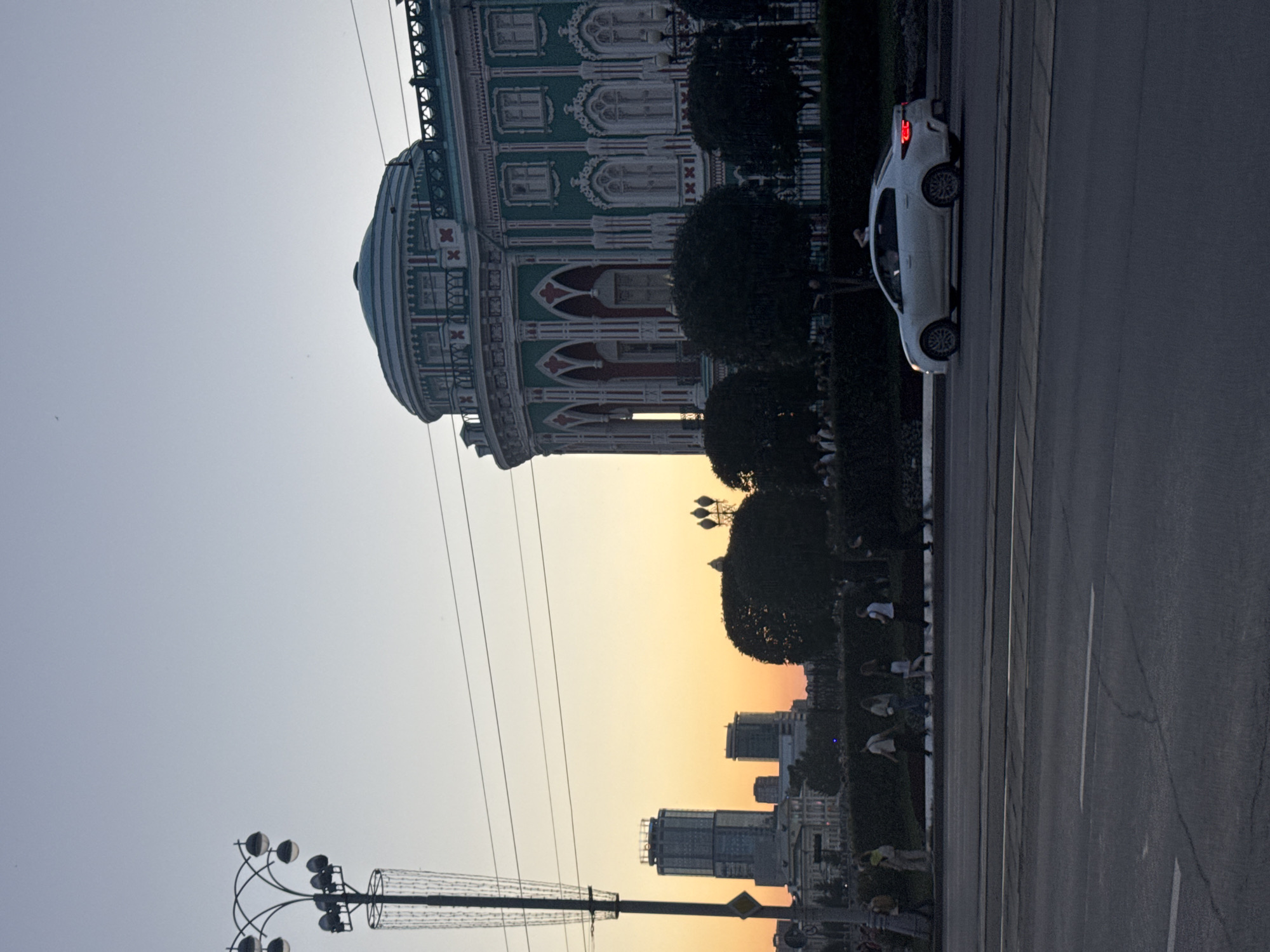 The image captures a street scene in Yekaterinburg, Russia, at dusk. In the foreground, a streetlamp with multiple illuminated bulbs stands on the left. A white sedan is driving on the right side of the road, its brake lights illuminated. Across the street, a line of people walks along a paved path. Behind them, trees and hedges form a dark silhouette against the fading light. In the background, the distinctive green and white facade of a large, ornate building with a domed roof is visible, likely the Uvarov Mansion or a similar historical structure. Further in the distance, modern skyscrapers punctuate the Yekaterinburg skyline, bathed in the warm hues of the sunset. The sky above is a gradient of pale blue and soft orange. Power lines stretch across the frame, indicating an urban environment.