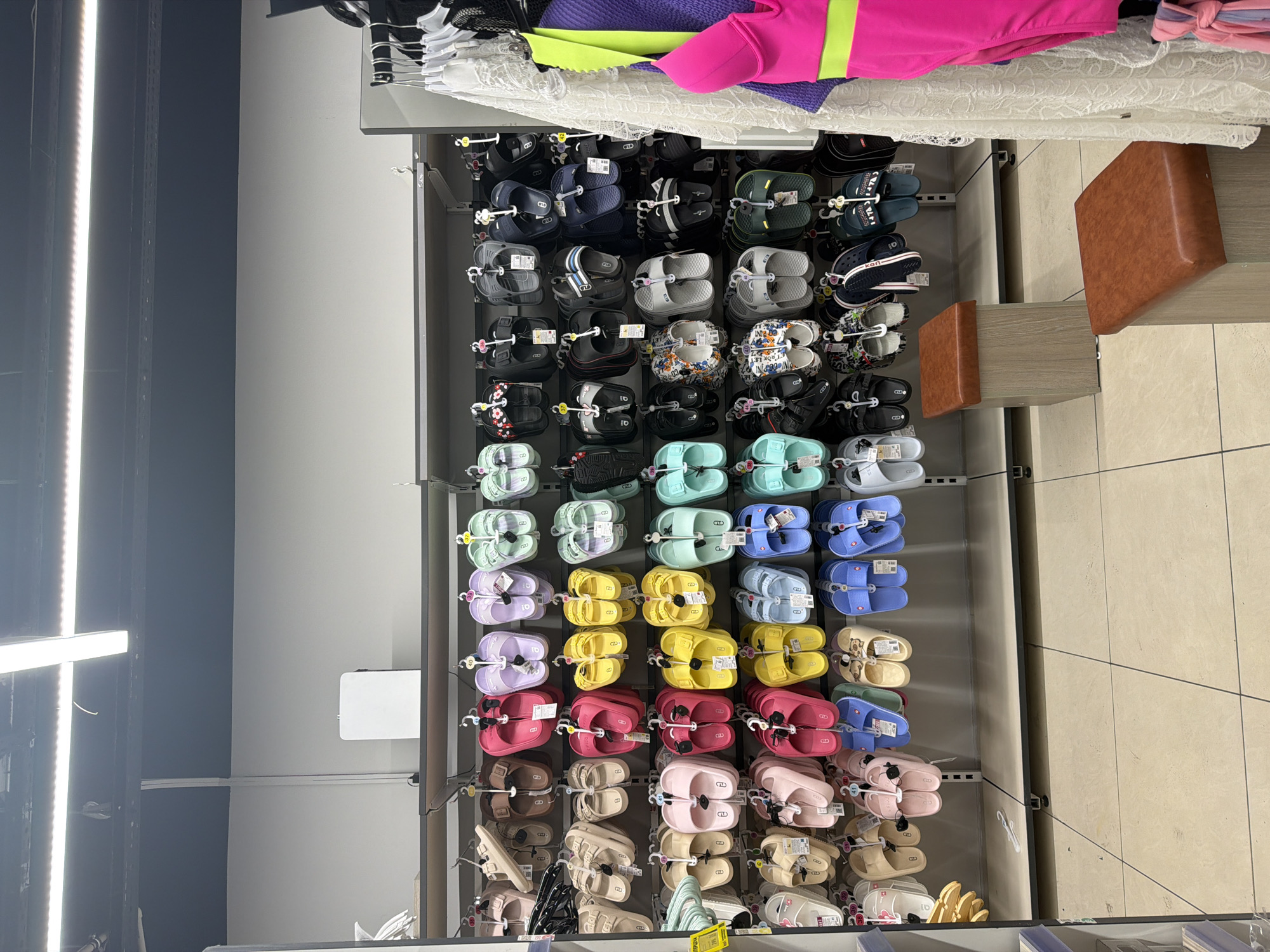 In Sochi, Russia, a retail store displays a wide assortment of flip-flops and slides on a wall-mounted shelving unit. The footwear, predominantly in bright colors like pink, yellow, purple, and blue, is organized in neat rows. Each pair is individually hung by its strap. The store environment is well-lit with overhead fluorescent lighting. Two small, wooden benches with brown cushioned tops are placed in front of the display, suggesting a fitting area for customers. To the right of the footwear display, a rack holds a few garments, including a white lace cover-up and a bright pink swimsuit. The flooring is tiled in a neutral beige color. No individuals are visible in the immediate scene.