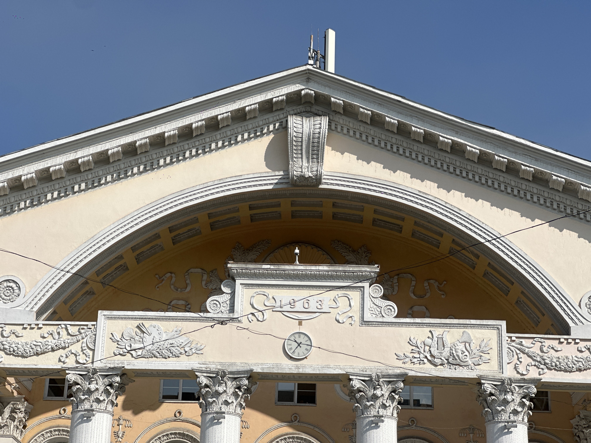 The image shows the facade of a building in Biysk, Russia. The building features classical architectural elements such as columns, ornate carvings, and a large arched structure above. A clock is visible on the facade, indicating the time. Above the clock, a year, "1903", is inscribed in a decorative frame. The sky is clear and blue, suggesting a sunny day. The building appears to be a significant public structure, possibly a theater or a historical institution, given its grandeur.