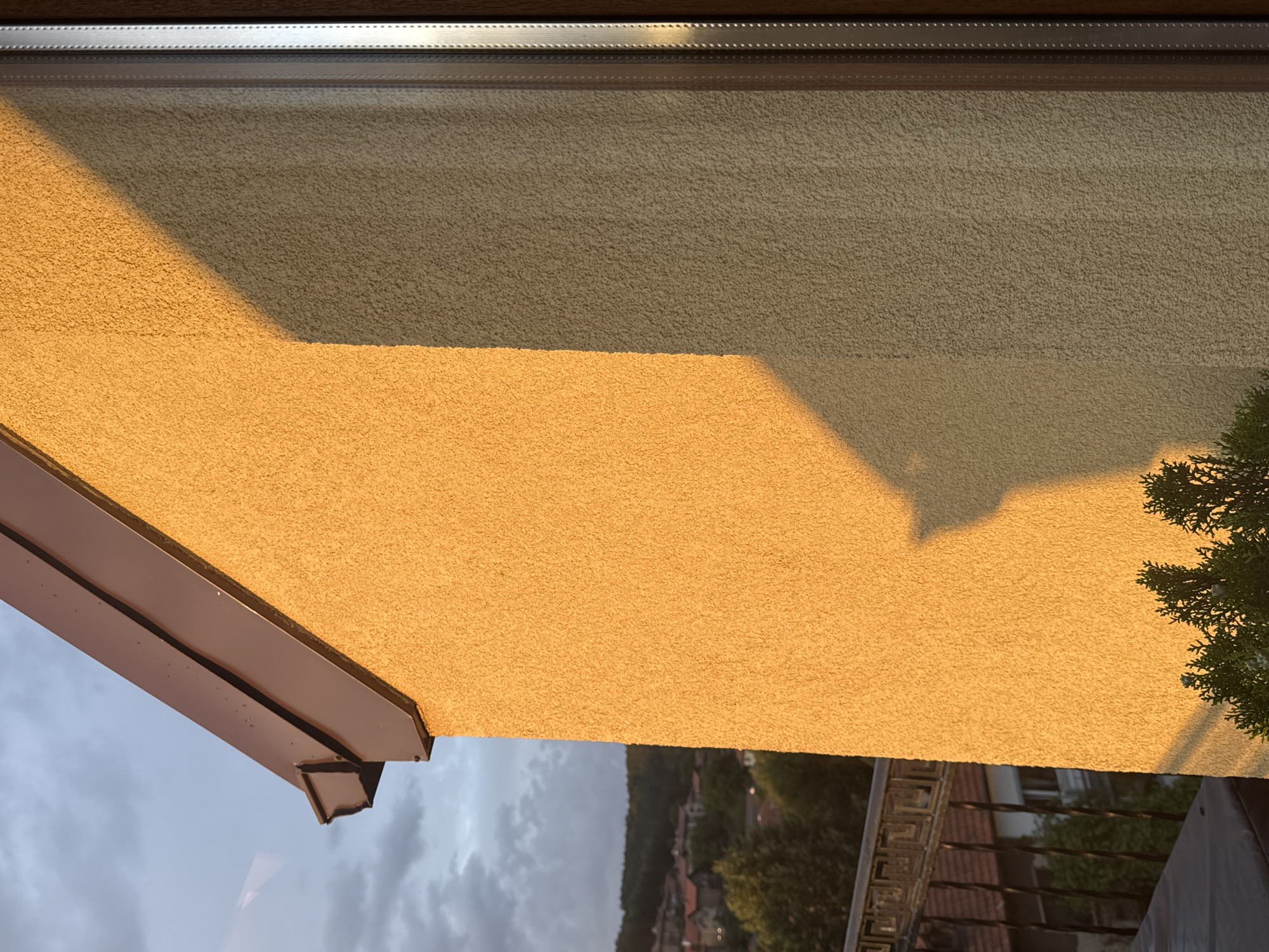 The image shows a close-up of a building's exterior wall, which is textured and painted in a light yellow color. A shadow, cast by an unseen object, stretches diagonally across the wall. A brown gutter runs along the roofline at the top left of the frame, with a section of the sky visible behind it. The sky is partly cloudy and appears to be either dawn or dusk due to the warm lighting.
To the left of the wall, a glimpse of a residential area is visible, with rooftops of houses nestled among trees on a hillside. A dark metal railing, part of a balcony or fence, is also present. In the foreground at the bottom, the dark green foliage of a small evergreen plant is visible. The right edge of the image features a window frame with vertical blinds.
While the image depicts an urban or suburban setting with a building and residential elements, there are no discernible clues within the image itself to pinpoint its specific location within the city of Pernik, Bulgaria. The general scene is a snapshot of an architectural detail against a natural and built environment backdrop, illuminated by natural light.