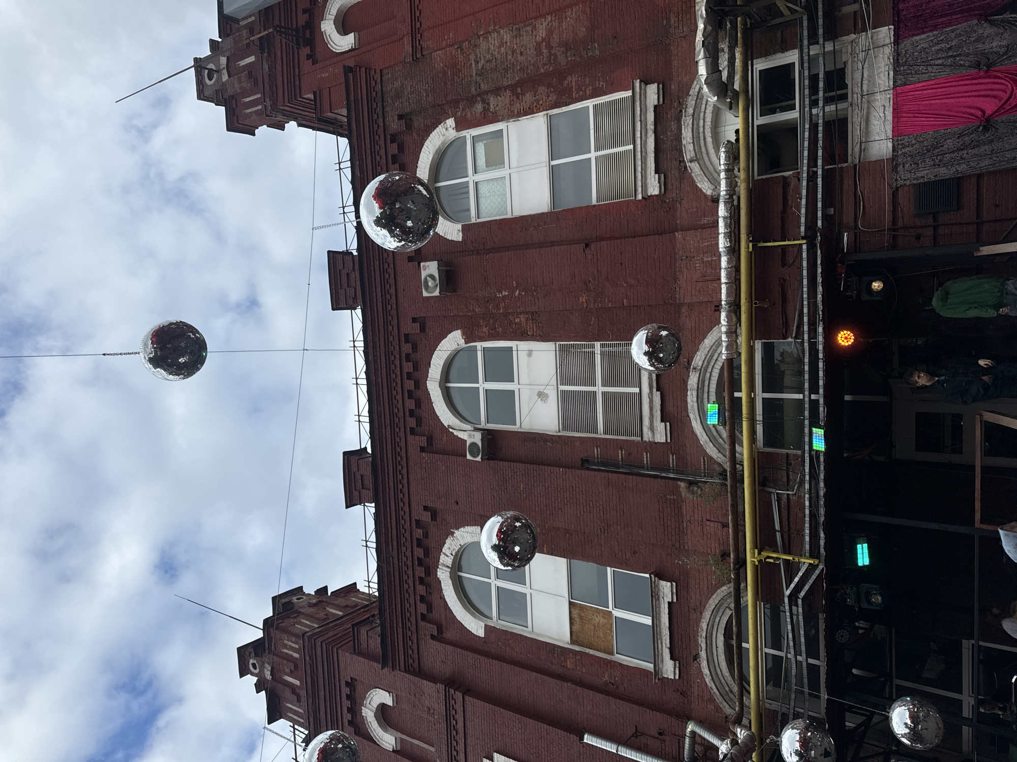 This is a ground-level, eye-level, medium shot of the exterior of a large, red brick building in Moscow, Russia. The building appears to be an old industrial or factory building that has been repurposed. The facade is characterized by arched windows and some modern additions such as ventilation pipes and air conditioning units. Several large, silver disco balls are suspended from the building by chains, indicating that it might be a venue for parties or events. Below the main facade, there is a darker area with some people visible, suggesting an entrance or a covered outdoor space. The sky above is cloudy. The image captures a unique blend of industrial architecture and festive decor.