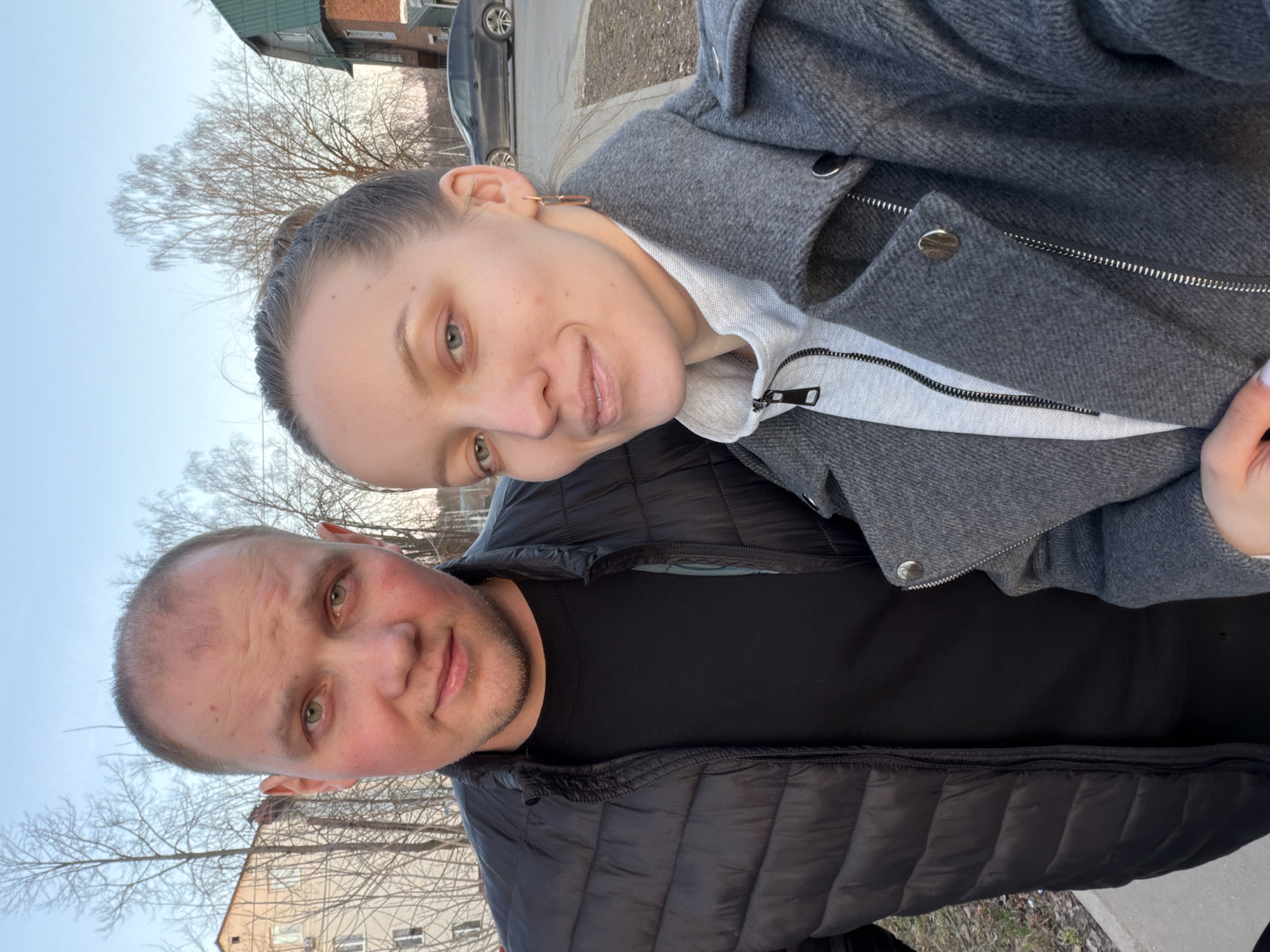 A selfie shows a man and a woman outdoors. The man is in the foreground, wearing a black puffer jacket and a black shirt. The woman is positioned behind him, wearing a gray wool coat over a white zippered sweater. Both are looking at the camera with slight smiles.
The background shows urban elements, including bare trees, buildings, a car, and a sidewalk. The photograph was taken in Reshetnikovo, Russia.