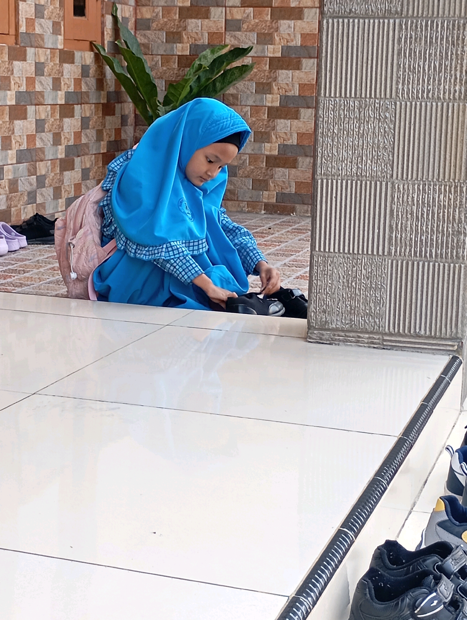 In Babelan, Indonesia, a young girl wearing a blue hijab and school uniform sits on a tiled floor. She is wearing a light pink backpack and is in the process of putting on her black shoes. To her left, two pairs of shoes are visible: one black and one light purple. The background consists of a textured brick wall and a decorative pillar. The floor is made of large, light-colored tiles. Several pairs of shoes are lined up on the right side of the image, suggesting a school or religious building setting.