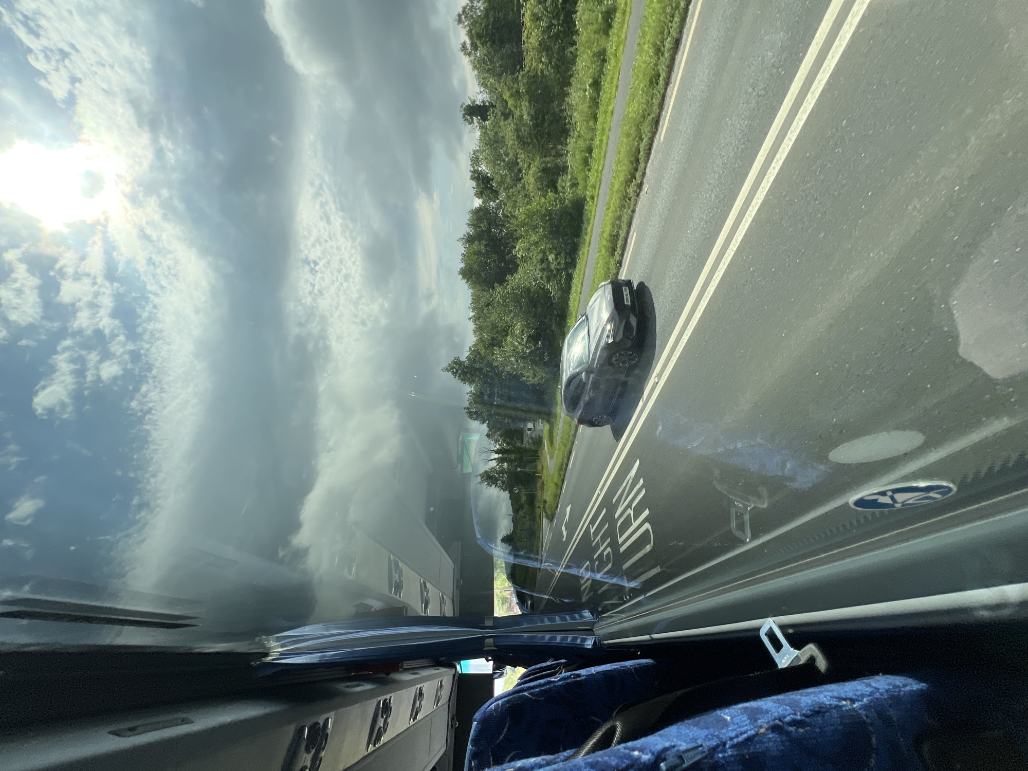 The image, taken from inside a vehicle, shows a road with trees and sky reflected on its surface. The scene appears to be a daytime shot in Letterkenny, Ireland. The weather seems to be partly cloudy, with sunlight breaking through the clouds.
The primary subjects are the road, the sky, and the surrounding vegetation. A dark-colored car is visible on the road, moving in the same direction as the viewer's vehicle. The road has double white lines marking the lanes. Reflections on the road surface reveal the sky, clouds, and trees, creating a mirrored effect.
Inside the vehicle, we see parts of seats covered in dark blue fabric with a pattern. A seatbelt buckle is visible, suggesting that the occupants are or were seated. The interior appears to be that of a bus or a large passenger vehicle.
Text is visible on the road surface. It appears to be a directional sign with white lettering. Partially visible words include "GHT" and "URN", possibly part of a place name or directional instruction. There is also a circular blue and white logo visible on the side of the vehicle.
The overall impression is a snapshot of travel on a highway, with a focus on the interplay between the natural environment and the man-made road. The reflection of the sky and clouds adds a dynamic and somewhat ethereal quality to the image. The time of day is clearly daytime, indicated by the bright sunlight.