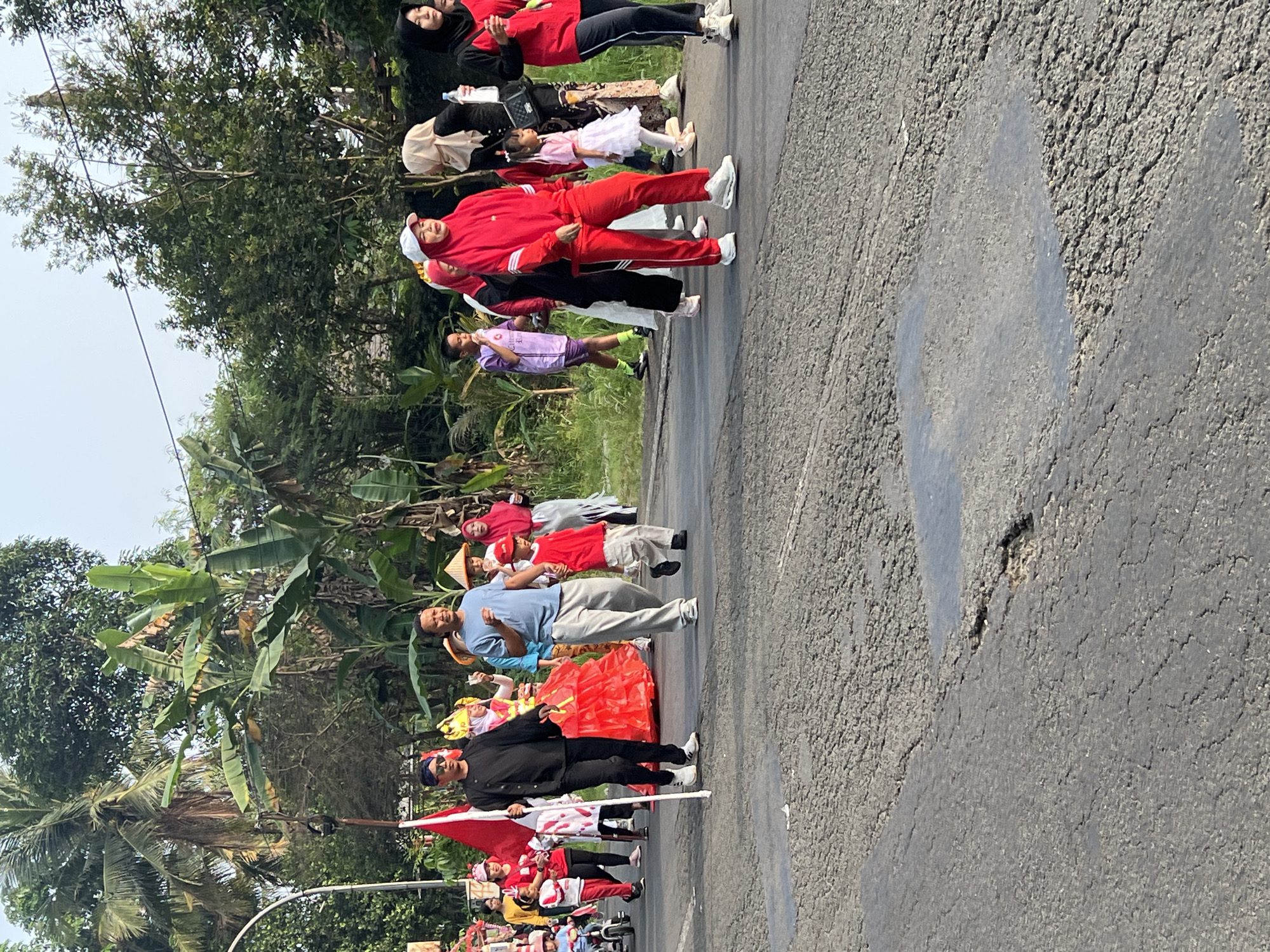 This is a medium-shot, eye-level, outdoor photograph of a parade or procession taking place on a paved street in Cibinong, Indonesia. The scene is populated by a diverse group of people, including adults and children, many of whom are dressed in celebratory attire.
In the foreground, a man in a black outfit with a blue head covering and sunglasses holds a white flagpole with a red and white flag. He is walking on the asphalt road, which shows signs of wear, including cracks and puddles. To his right, a young boy in a red shirt and beige pants walks alongside a man in a light blue t-shirt and light gray pants.
Further into the procession, several individuals are visible. There is a person in a bright red dress with elaborate headwear, and another person in a red tracksuit and white cap. Other participants are seen in various colored clothing, including purple, white, and black. Some individuals are wearing hijabs. A child in a pink dress and white shoes is also present.
The background features lush green trees, including palm trees and banana trees, suggesting a tropical or subtropical environment. Overhead, a power line is visible against a clear sky. The overall impression is of a community event or cultural celebration.