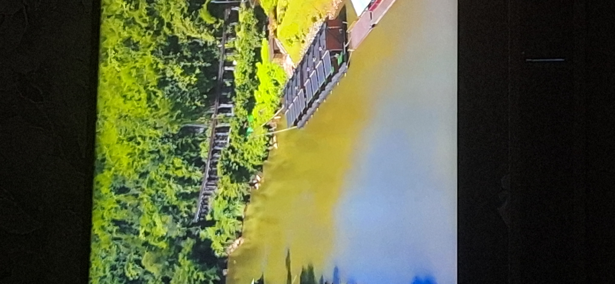 The image depicts a body of water, likely a river, with lush green foliage along its banks. In the foreground, a series of dark, rectangular structures are floating on the water, appearing to be some form of raft or barge system. These structures have a segmented appearance with visible divisions. Further in the background, a long, elevated structure with numerous vertical supports can be seen stretching across the landscape, partially obscured by the dense greenery. This structure resembles a bridge or an old railway line. The water itself has a yellowish-brown hue and reflects the surrounding environment. The overall scene suggests a natural landscape with human-made elements integrated into it. There is no identifiable content that relates to the city of Bila Tserkva, Ukraine.