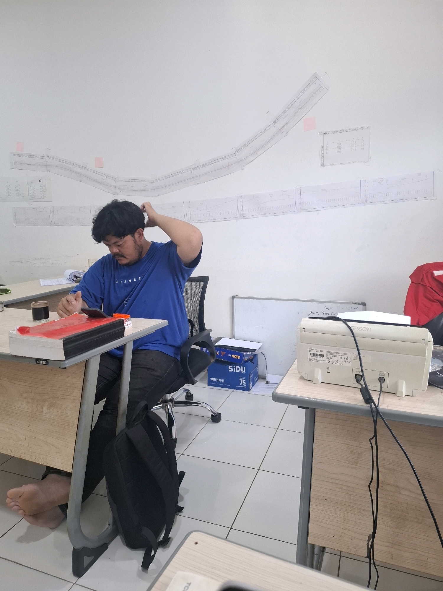 In an office space in Kutasirna, Indonesia, a man with dark, curly hair and a mustache, wearing a blue t-shirt and black pants, is seated at a desk. He is scratching his head with his right hand and looking down at his phone, which rests on a red object on the desk. To his left, a glass of dark liquid, likely coffee, sits on the desk next to a black binder. A black backpack is visible on the floor behind the man, attached to his office chair. The walls are adorned with large blueprints and smaller sheets of paper, suggesting an architectural or engineering context. To the right of the man, another desk holds a white printer and some boxes of paper. The floor is tiled in a white grid pattern.