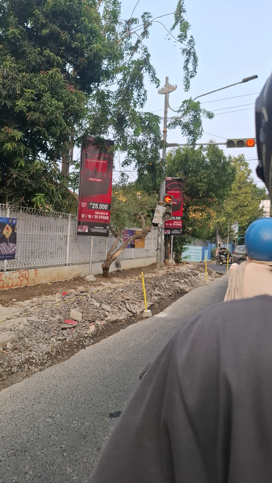 This image depicts a street scene in Bandar Lampung, Indonesia, likely during daylight hours given the lighting. The foreground shows the perspective from the back of a motorcycle, with a dark grey garment partially obscuring the view. The road surface is asphalt. To the left, a construction area is visible with loose gravel, rubble, and some red debris, bordered by a metal fence. Yellow poles are present in the construction zone.
Behind the fence, several tall trees with green foliage are present. Two vertical banners are prominently displayed. The banner on the left is black with red accents and displays text including "FILTER BLACK" and "MOTION," along with a price of "RP 25.000" and availability information. The banner on the right is similar in design and content.
Further down the street, traffic lights are visible, with the red light illuminated. Another banner of a similar design is located near the traffic light pole. In the background, more trees and greenery can be seen, along with a clear sky. A few vehicles, including a motorcycle with riders, are visible further down the road. The overall impression is a street undergoing some form of development or maintenance, with commercial advertisements present.