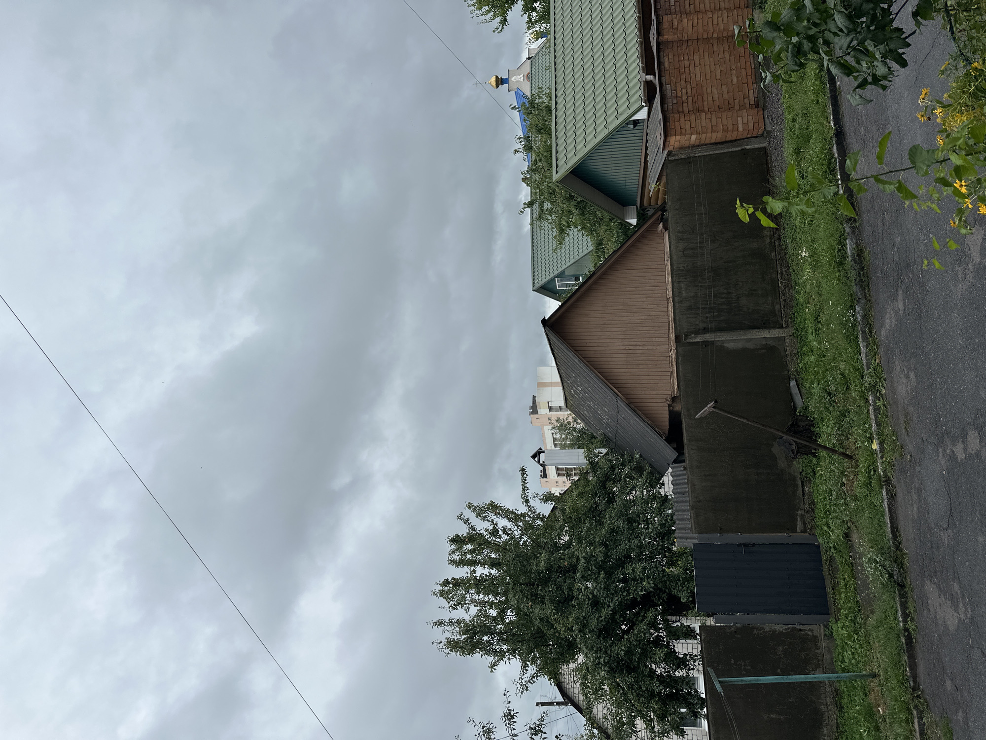 The image depicts a residential area under a cloudy sky in Khmilnyk, Ukraine. In the foreground, a paved road with some cracks runs horizontally, bordered by a strip of green grass and vegetation. A dark grey metal gate is visible in the mid-ground, set within a dark concrete fence. Behind the fence, several houses with pitched roofs are visible, some with green metal roofing and others with brown wooden siding. A large green tree stands to the left of the gate, and a taller modern building with light-colored facades and windows is visible in the background behind the houses. To the far right, a golden dome and a blue roof of a church or religious building are partially visible. The overall atmosphere is subdued, consistent with overcast weather. No people are present in the scene.