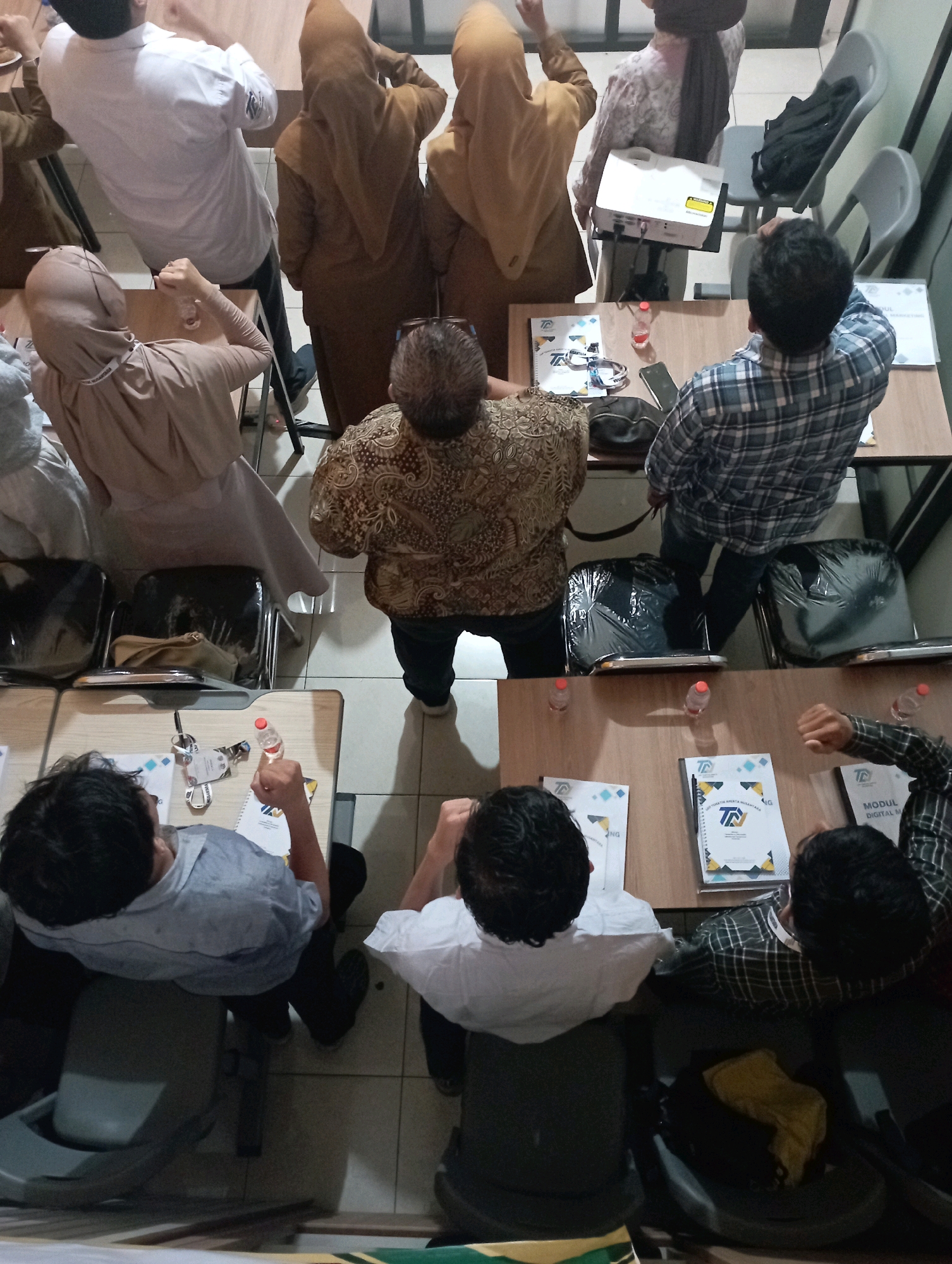 In a well-lit indoor space, likely a classroom or conference room, a group of individuals are engaged in a meeting or presentation. The scene is viewed from a high angle, looking down on the participants and the tables. The room features beige tiled flooring and several tables arranged in rows.
In the foreground, two men are seated at a table, facing away from the camera. One man, wearing a light blue collared shirt, is looking down at the table. The other man, in a plaid shirt, has his arms raised and fists clenched, appearing to be actively participating or demonstrating something. A bottle of water sits on the table in front of him. Notebooks with logos, identified as "T" and text such as "DIGITAL M" and "MODUL," are laid out on the table, suggesting a training or educational setting.
Further back, a man in a patterned brown shirt is standing, his back to the camera, and he appears to be gesturing with his hands. Several women in brown hijabs are seated or standing around other tables, some looking towards a projector on a table. One woman in a light brown hijab is seated, looking towards the front. Another woman, also in a light brown hijab, is seated and looking down.
A white projector is placed on a table, with a black bag next to it. Several chairs, some covered in black plastic, are visible throughout the room. The overall atmosphere suggests a formal gathering, possibly a workshop or seminar, taking place in Tasikmalaya, Indonesia, given the visual context and cultural attire.
