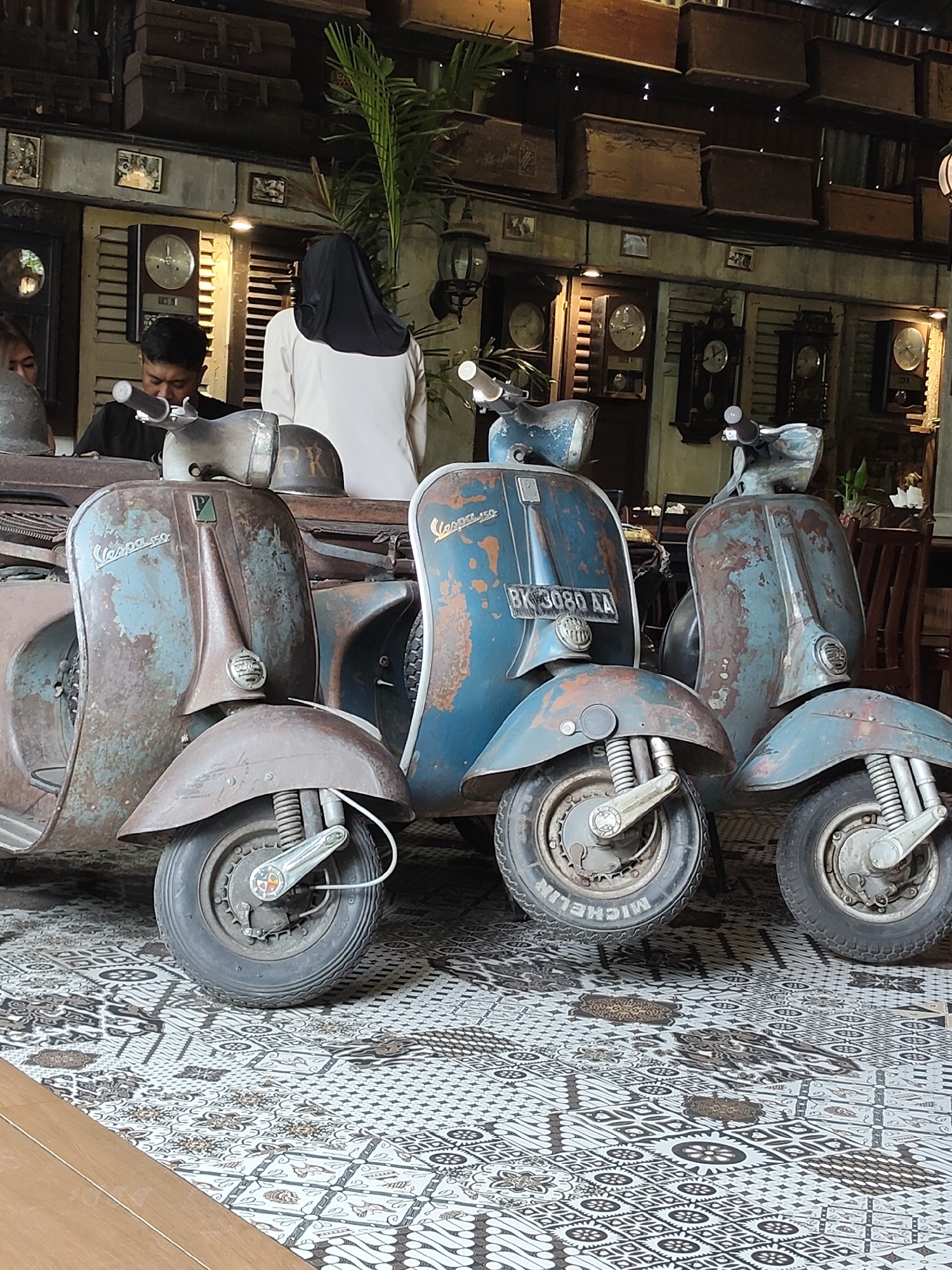 This image captures a row of three weathered, vintage Vespa scooters parked on a patterned tiled floor. The scooters exhibit signs of age, with rust and faded paint. The leftmost scooter is predominantly brown with a light blue accent, while the middle and rightmost scooters are a faded blue with significant rust patches. The middle scooter has a license plate that reads "BK 3080 AA."
Behind the scooters, the environment suggests an indoor setting with a rustic, antique aesthetic. The background features wooden shelves stacked with what appear to be old boxes or suitcases. Several vintage clocks are mounted on a wall, contributing to the retro ambiance. A person wearing a black headscarf and a light-colored garment stands behind the middle scooter, facing away from the camera. Another person, a man wearing a black shirt, is partially visible to the left, appearing to be looking down. The floor is covered with a black and white patterned tile, possibly batik. This scene is likely located in a vintage-themed cafe or shop in the city of Medan, Indonesia, given the style of the scooters and the general decor.