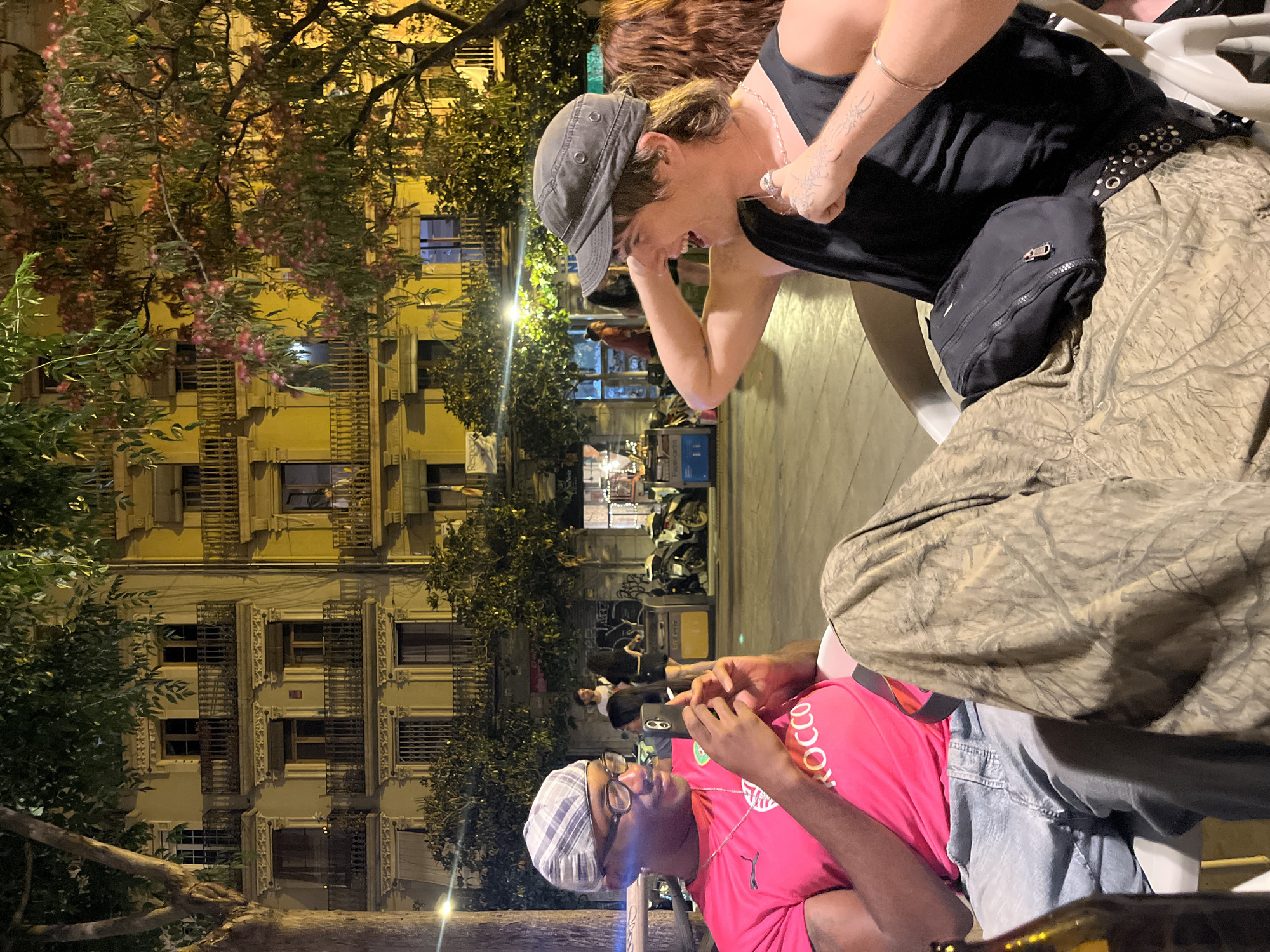A street scene in Barcelona, Spain, at night. Two individuals are seated in outdoor chairs. One person, wearing a pink shirt and glasses, is holding a smartphone. The other individual, wearing a black tank top and a grey cap, is leaning forward. Buildings with wrought-iron balconies are visible in the background. Trees with pink blossoms are also visible. Motor scooters are parked along the street.