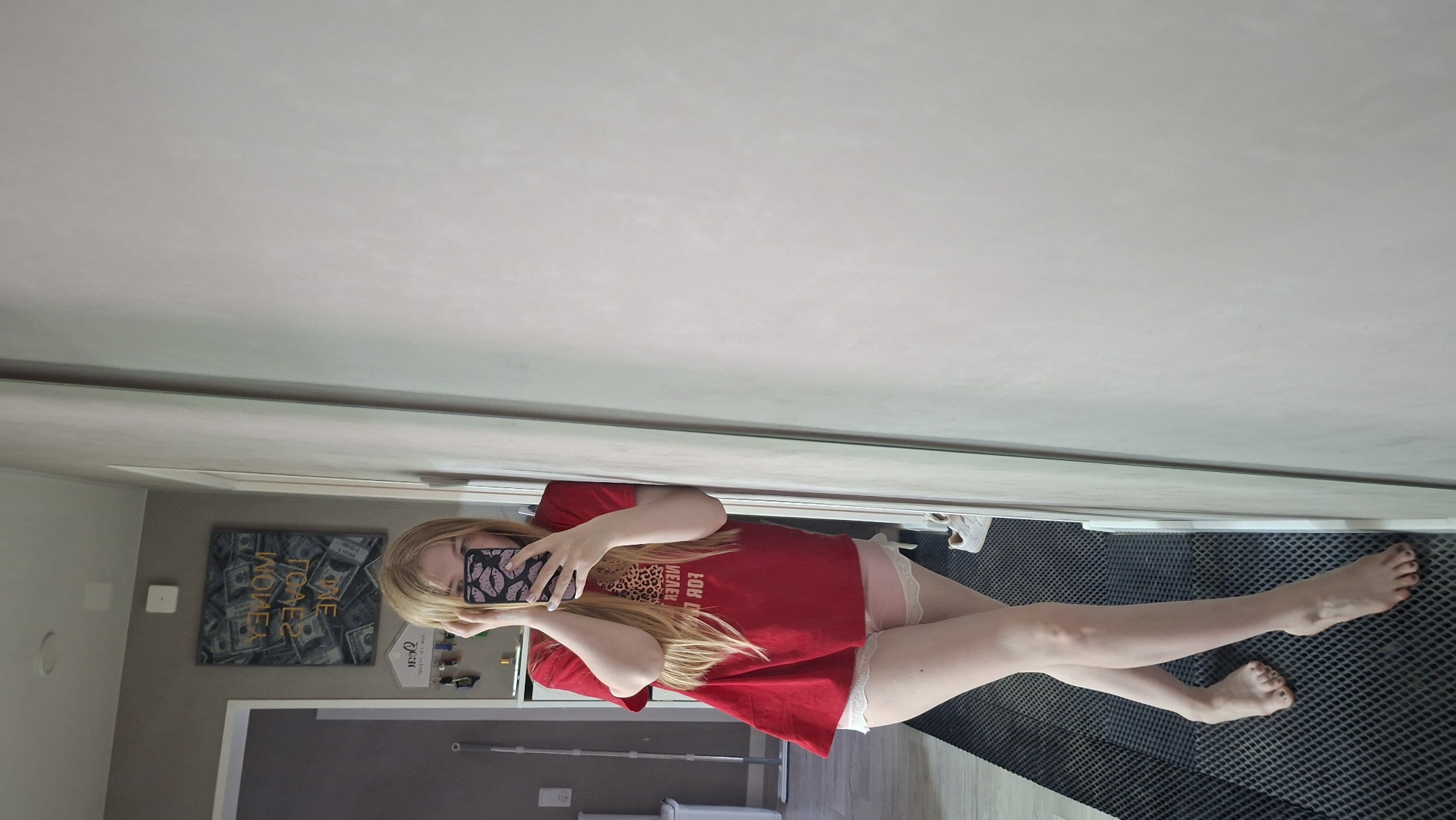 A blonde woman is lying on the floor, taking a photo of herself with her phone. She is wearing a red t-shirt and pale pink shorts with white lace trim. Her legs are extended outward, and her bare feet are visible. The floor is covered with a dark grey, textured mat.
In the background, there is a wall with a picture of money and a white cabinet with various items. A mop or broom is leaning against the wall near a white object, possibly a toilet. A white switch is also visible on the wall. The location is likely an indoor space, such as a home or apartment. There is no information to indicate that the scene is in Chaikovsky, Russia.