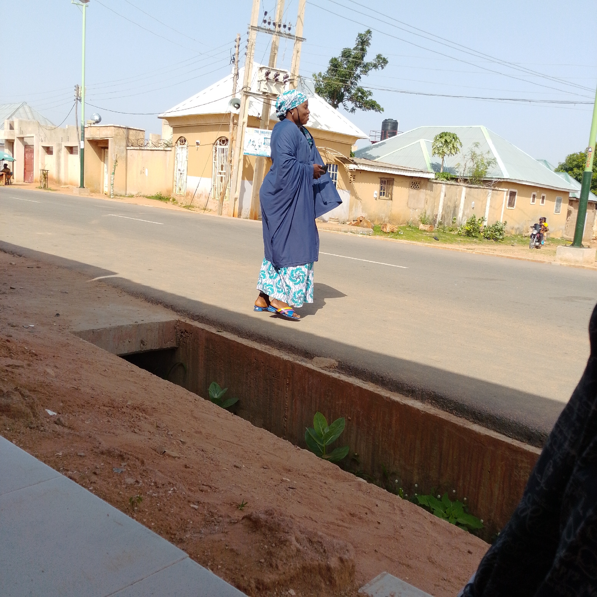 An eye-level, medium shot captures a woman walking along the side of a paved road in Gombe, Nigeria. The woman, dressed in a dark blue abaya and a blue and white patterned headscarf, walks away from the camera. She wears multi-colored sandals. Her floral skirt is visible beneath the abaya. The road has a white line marking the center. On the left of the road, a drainage ditch runs parallel to it, filled with dirt and some green plants. Behind the woman, a row of beige buildings with white roofs lines the street. A tree with sparse green leaves is visible behind the buildings. In the distance, a person rides a motorcycle. The sky is clear and pale blue.